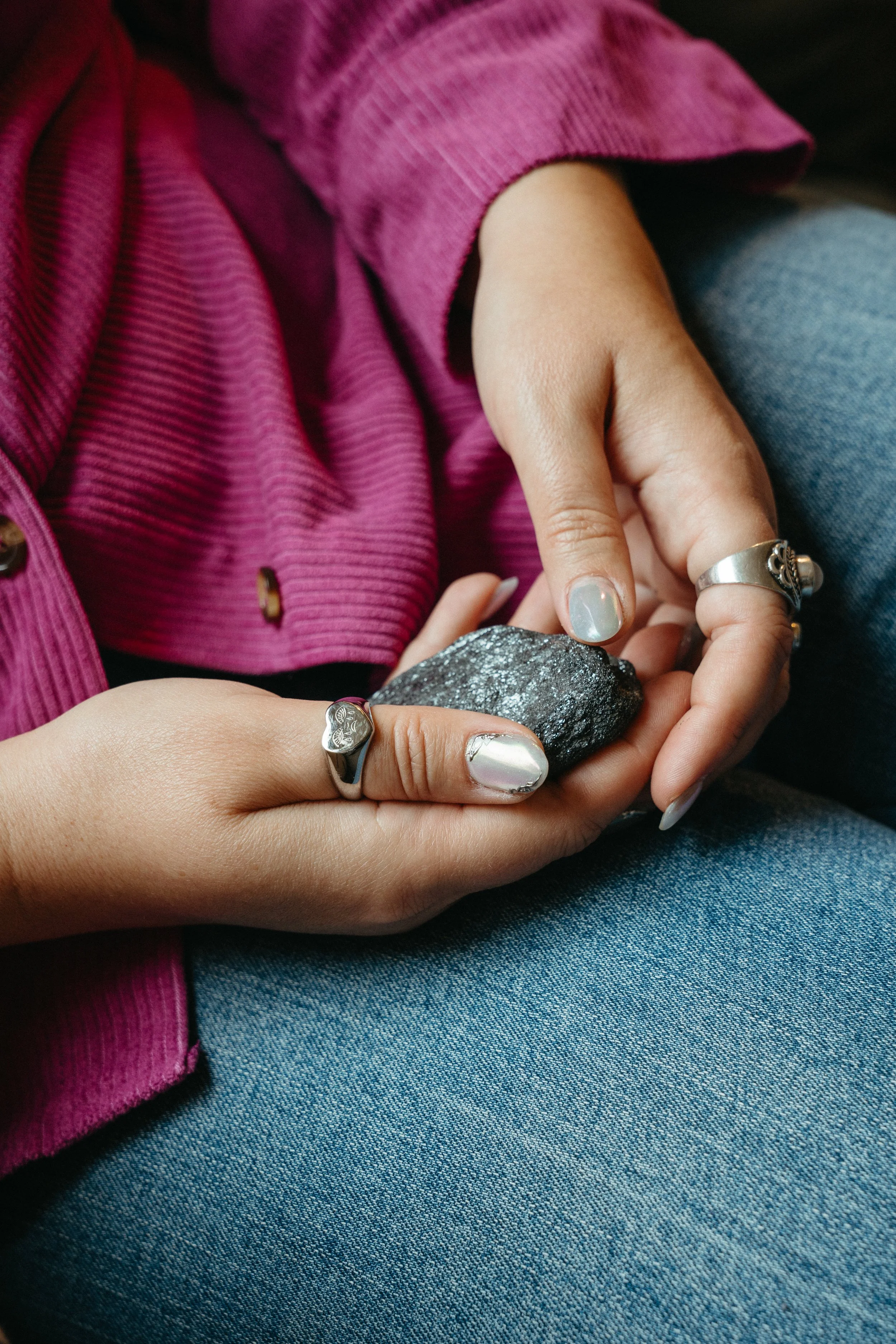 A photo of two hands with pearlescent nails and silver rings holding a gray rock in a lap