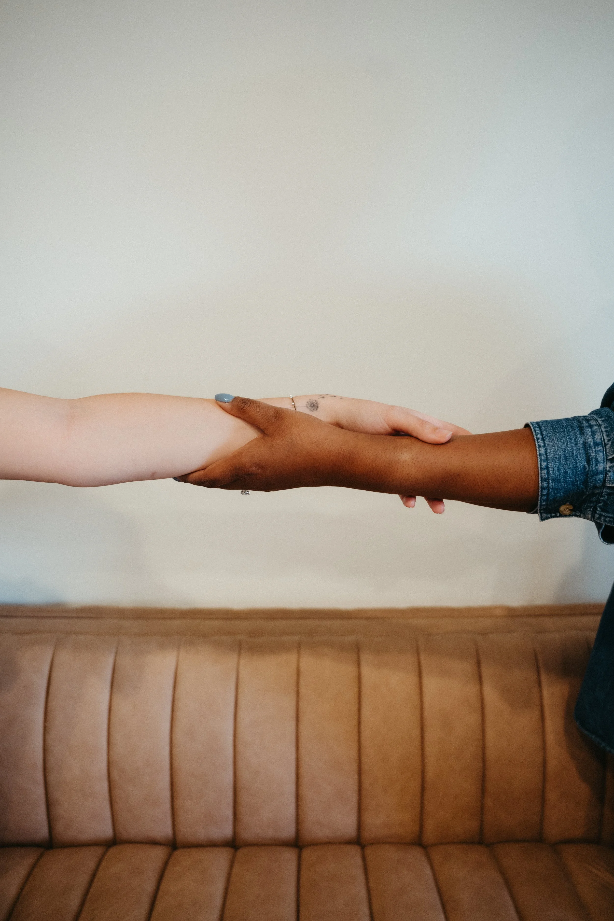 Photo of the arm of a white person with tattoos clasping the arm of a Black person with blue nails and a jean jacket