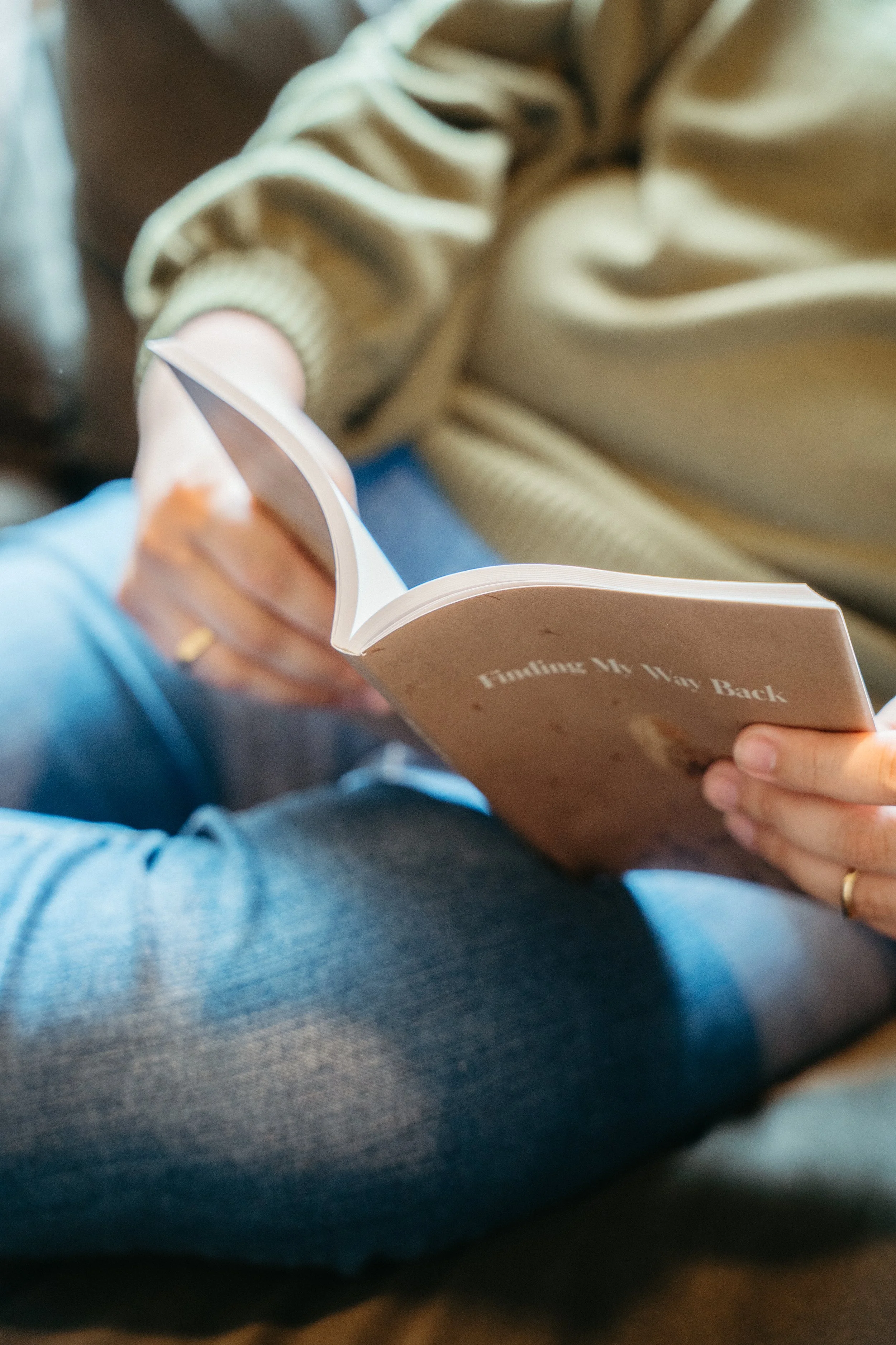Photo of a person in a cozy green sweater and jeans reading a paperback book called Finding My Way Back