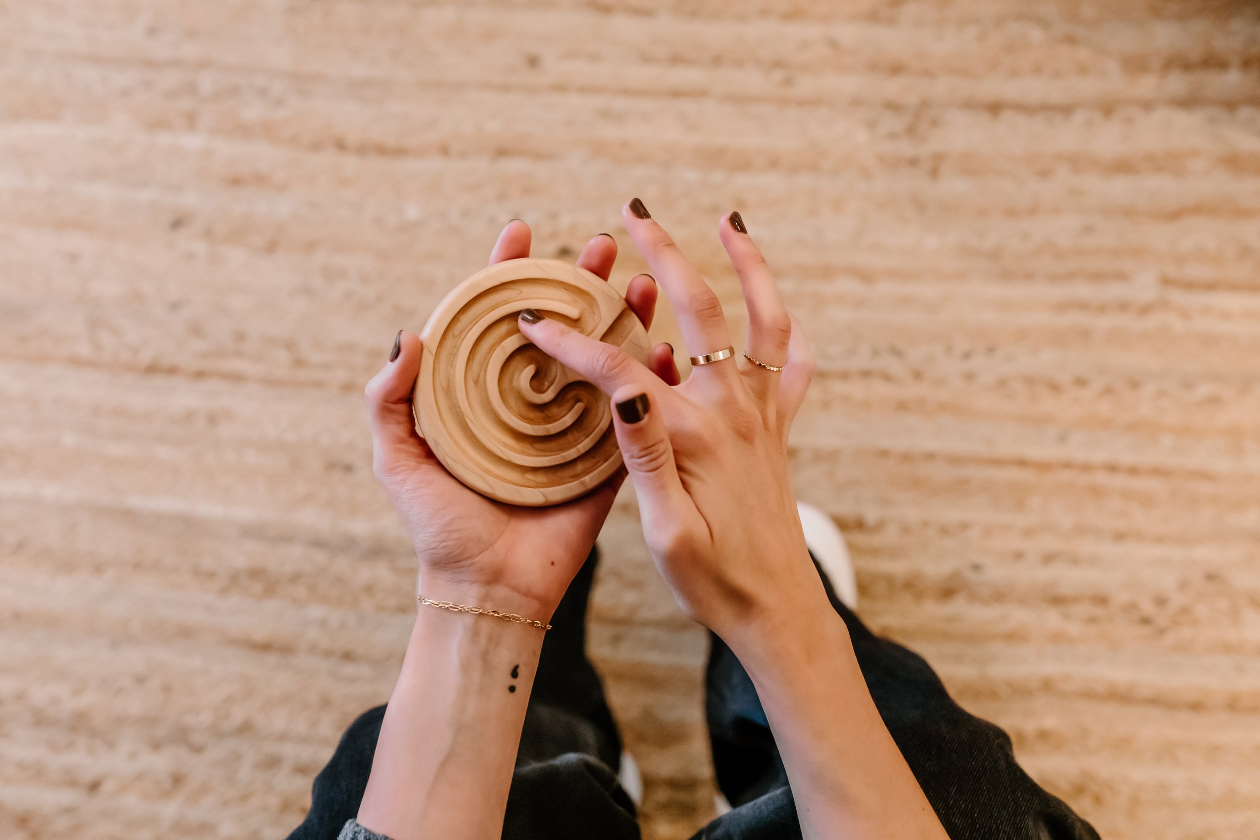 A photo of a hand with dark chocolate nails and gold rings touches a small wooden coaster that looks like a circular maze