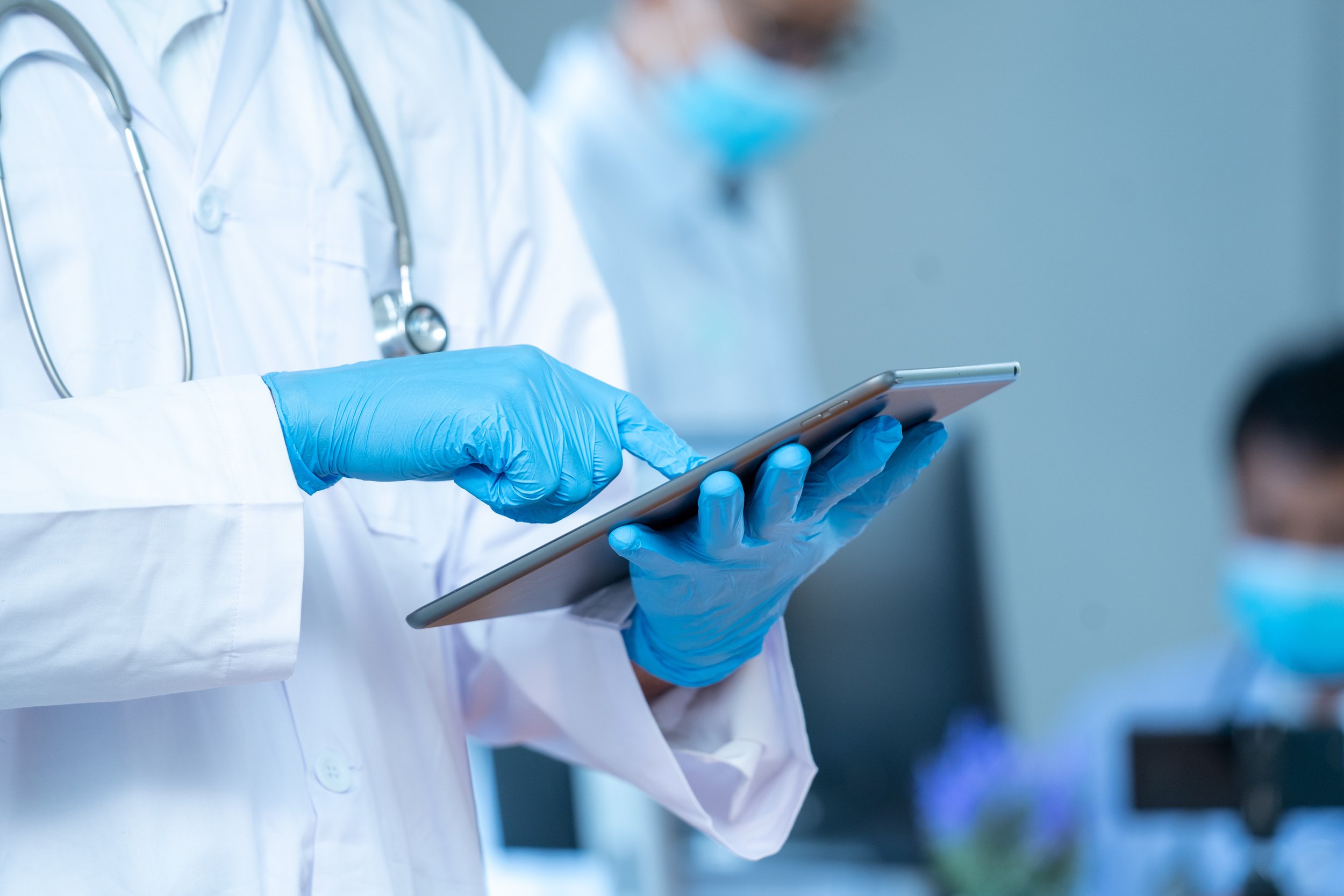 A medical professional wearing a white coat, blue gloves, and a stethoscope around their neck holding a tablet device, with another person blurred in the background.