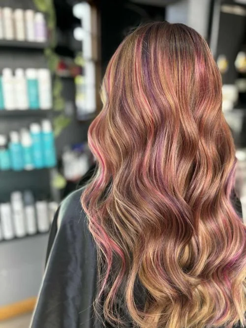 Back view of a woman with long, wavy hair featuring shades of pink, blonde, and brown in a salon setting.