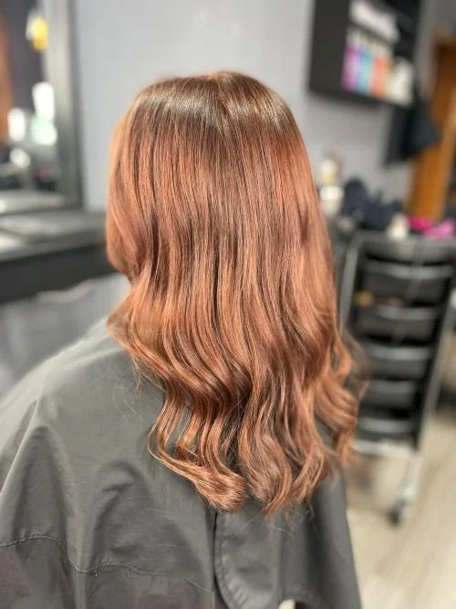 Back view of a woman with medium-length, wavy, reddish-brown hair in a salon.