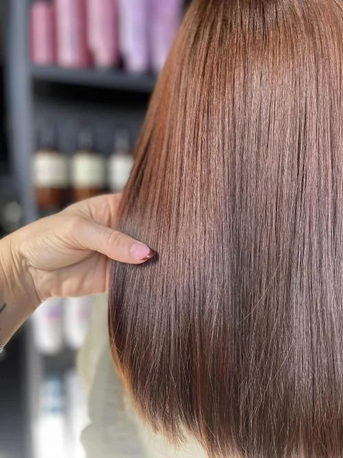 Close-up of a person holding a section of shiny, straight, reddish-brown hair against a blurred background of hair care products.