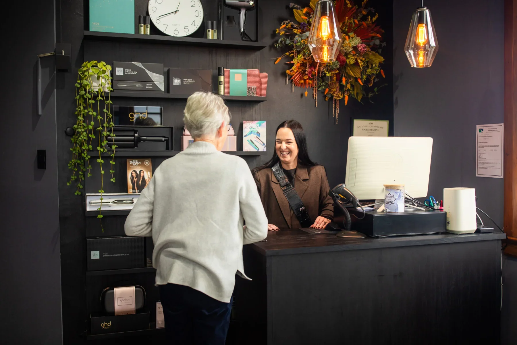 A customer and receptionist smiling and talking at a reception desk in an office or salon. Behind the receptionist are shelves with various items, a clock, and a colorful floral arrangement. The reception area has modern lighting and decor.