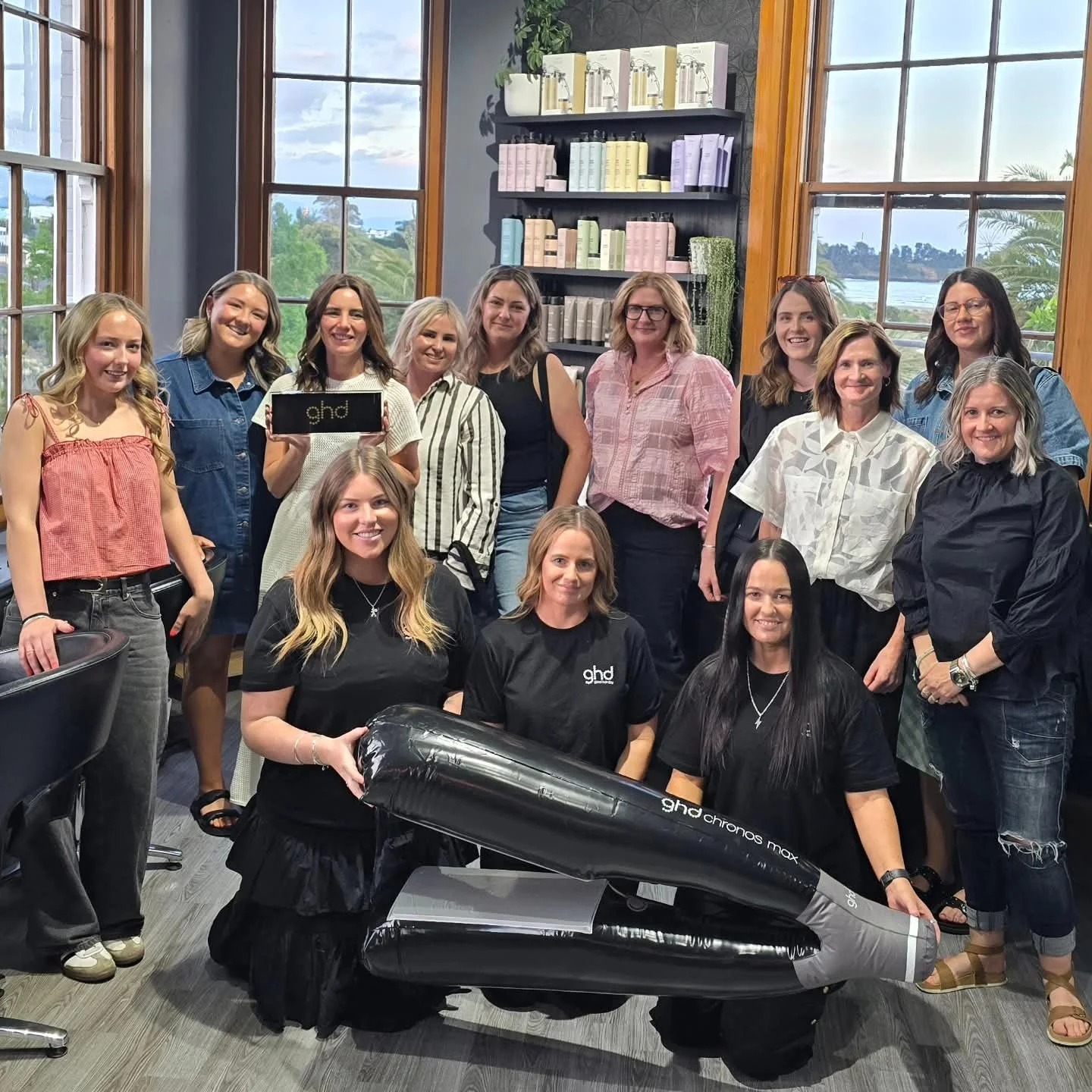Group of women gathered indoors for a photo, some holding a sign that says 'ghd' and an inflatable hairdryer prop, in front of shelves with hair and beauty products, large windows showing a green landscape outside.