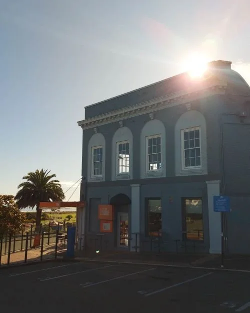 A blue two-story building with arched windows and decorative trim, set against a bright sky with the sun partially obscured, and a palm tree in the background.