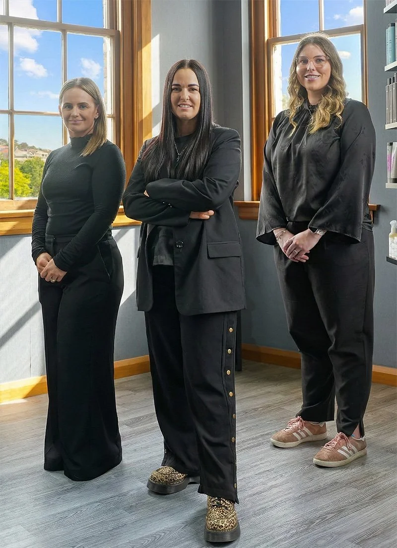 Three women standing inside a room with large windows, dressed in black clothing and smiling at the camera.