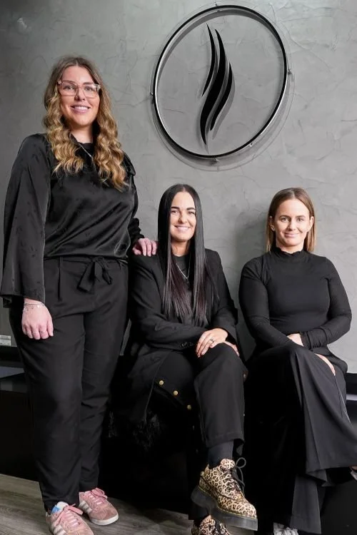 Three women dressed in black clothing, two sitting and one standing, in front of a gray wall with a circular logo featuring three black wavy lines.