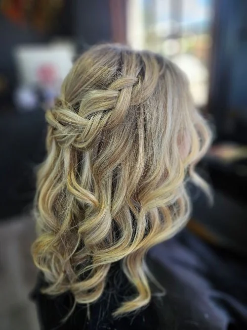 Woman with blonde, curly hair styled with a side braid and loose waves, sitting indoors near a window.