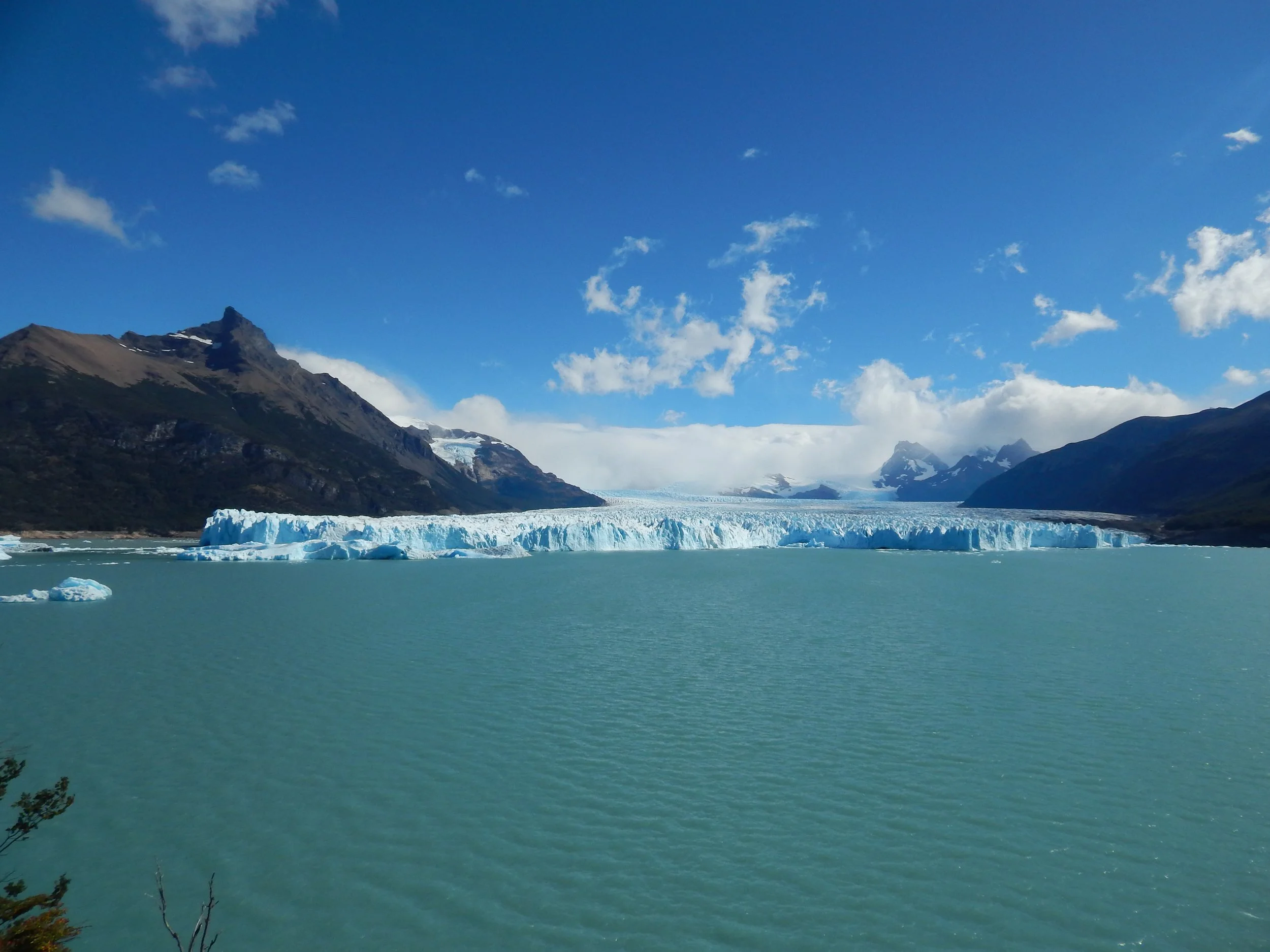 A vast glacier with jagged ice formations flowing into a turquoise lake, surrounded by snow-capped mountains under a partly cloudy blue sky.
