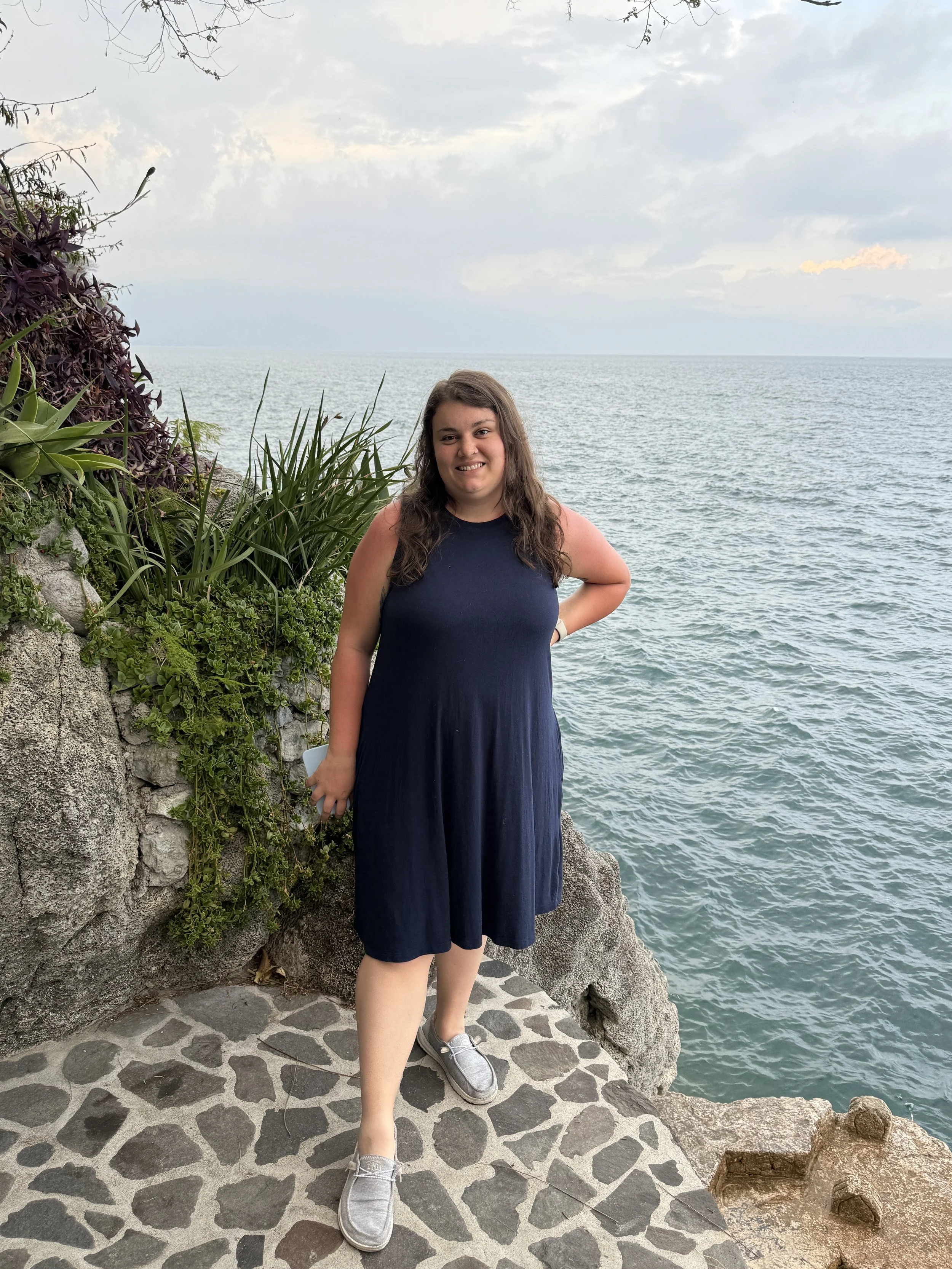 A young woman standing on a stone pathway near a blue lake, with greenery and rocks surrounding her, under a cloudy sky.