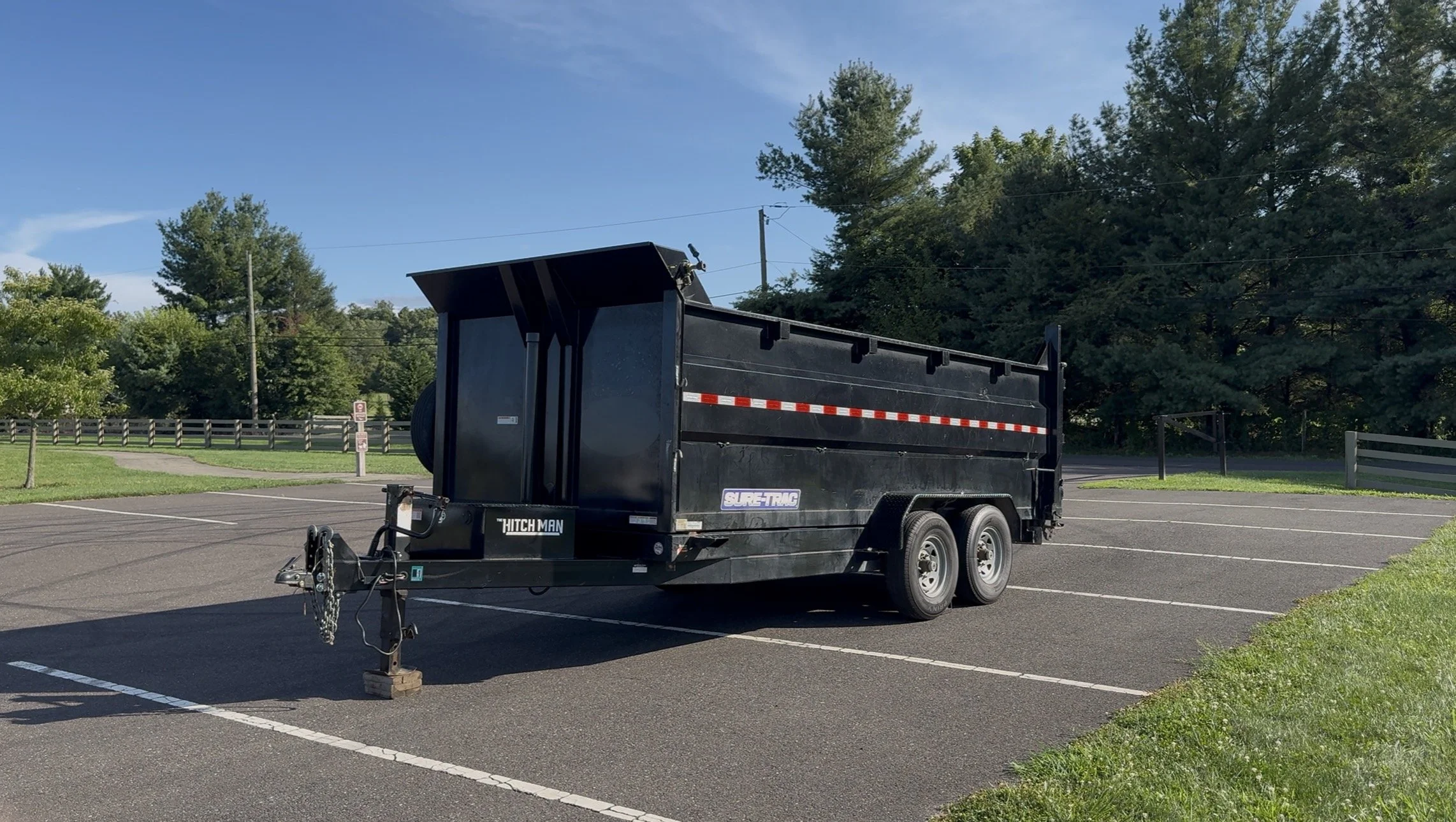 A black dump trailer parked in an empty parking lot with surrounding green trees and a blue sky.