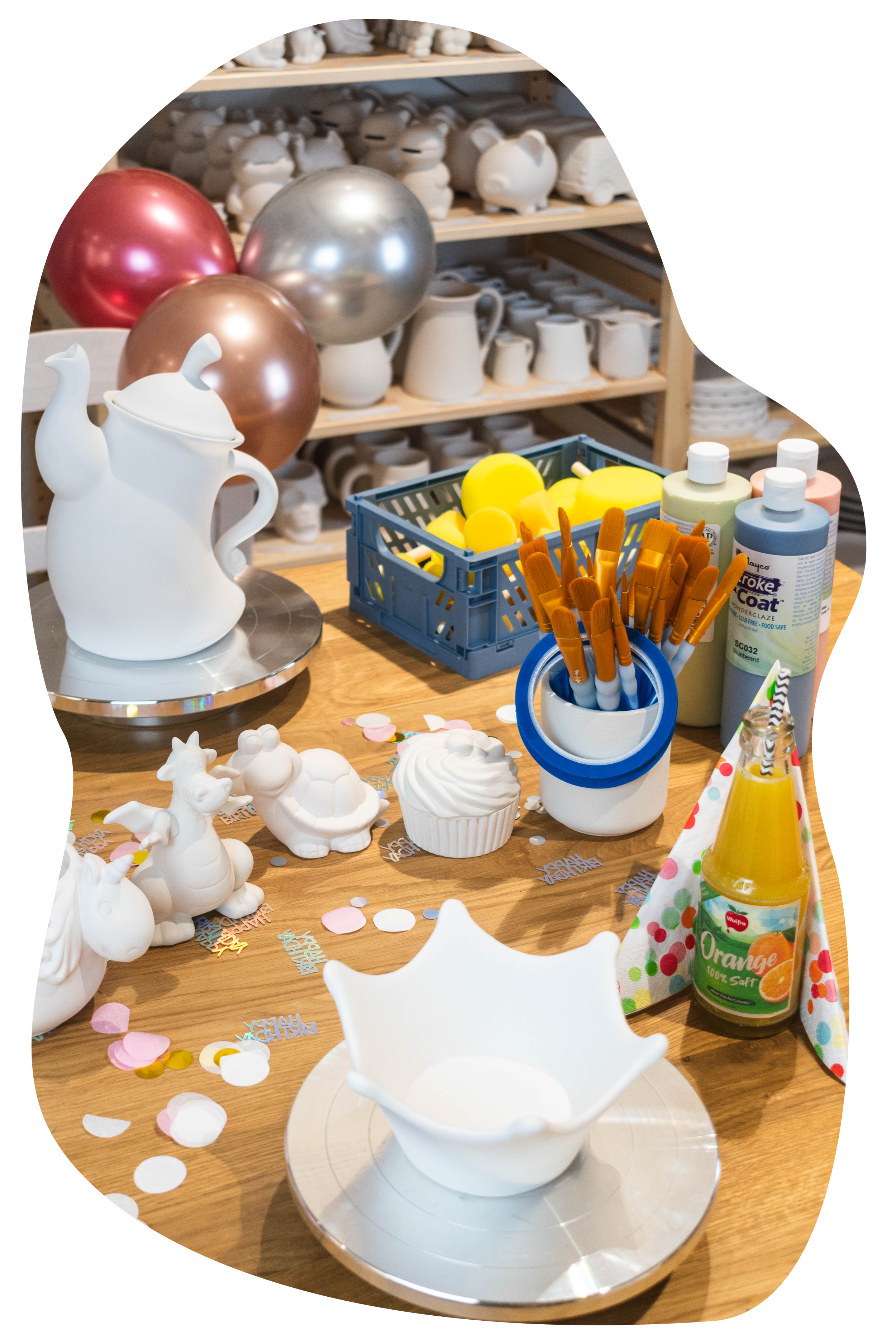 Basteltisch mit Cake Pops, Pinsel, Dekorationskonfetti, Knetmasse, Kerzen, und Glitzer in einem Raum mit mehreren Keramikfiguren auf Regalen im Hintergrund.