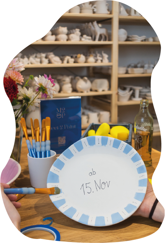 Person bemalt einen Pappteller mit blauen Akzenten, auf dem das Datum 15. Nov geschrieben ist, in einer kreative Werkstatt mit Pinsel und Farben. Im Hintergrund sind Regale mit unbemalten Keramikstücken, und im Vordergrund stehen Blumen, Pinsel, Schwämme und eine Glasflasche auf dem Tisch.
