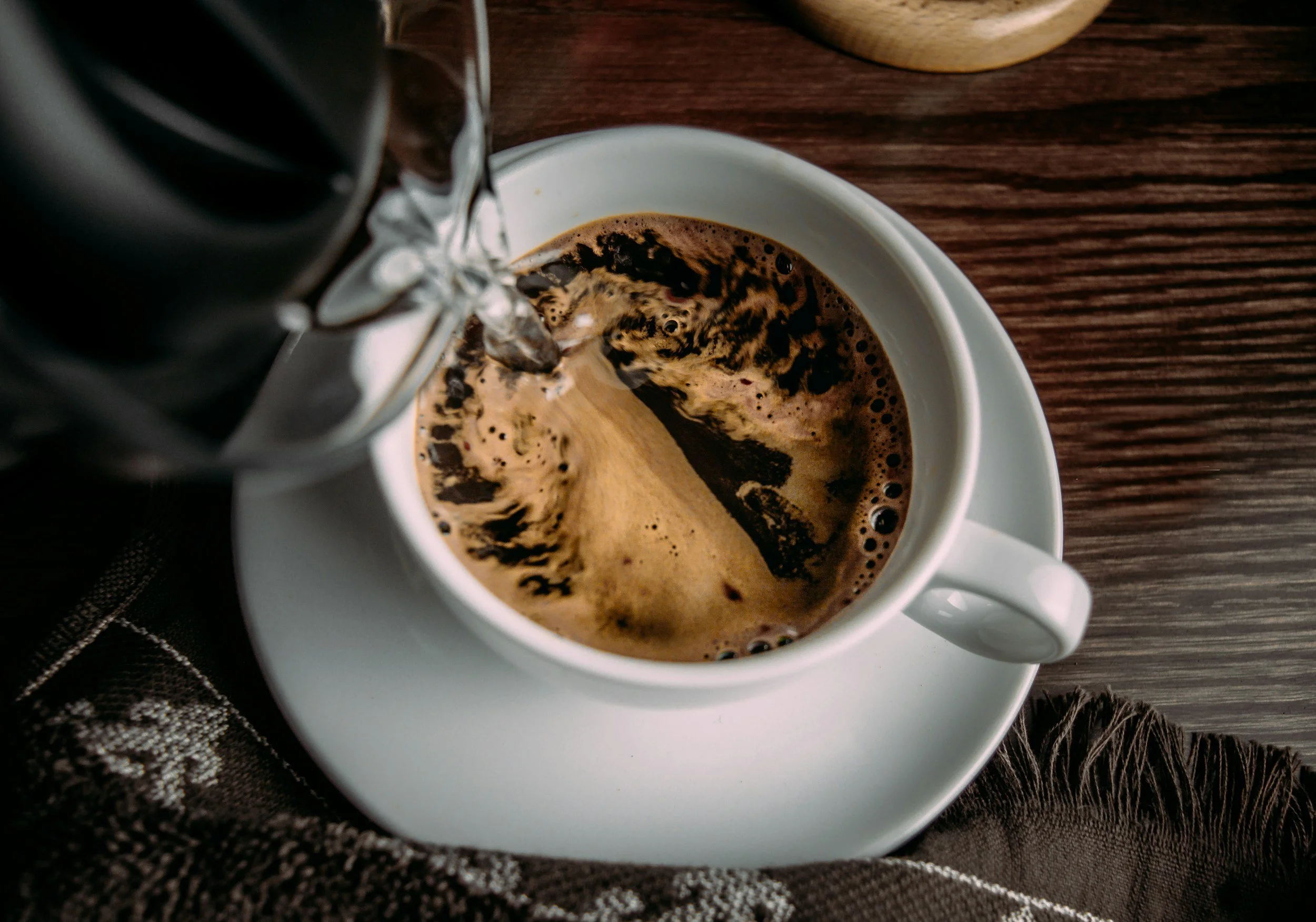A cup of hot coffee with dark foam swirling inside, with hot water being poured into it from a kettle.