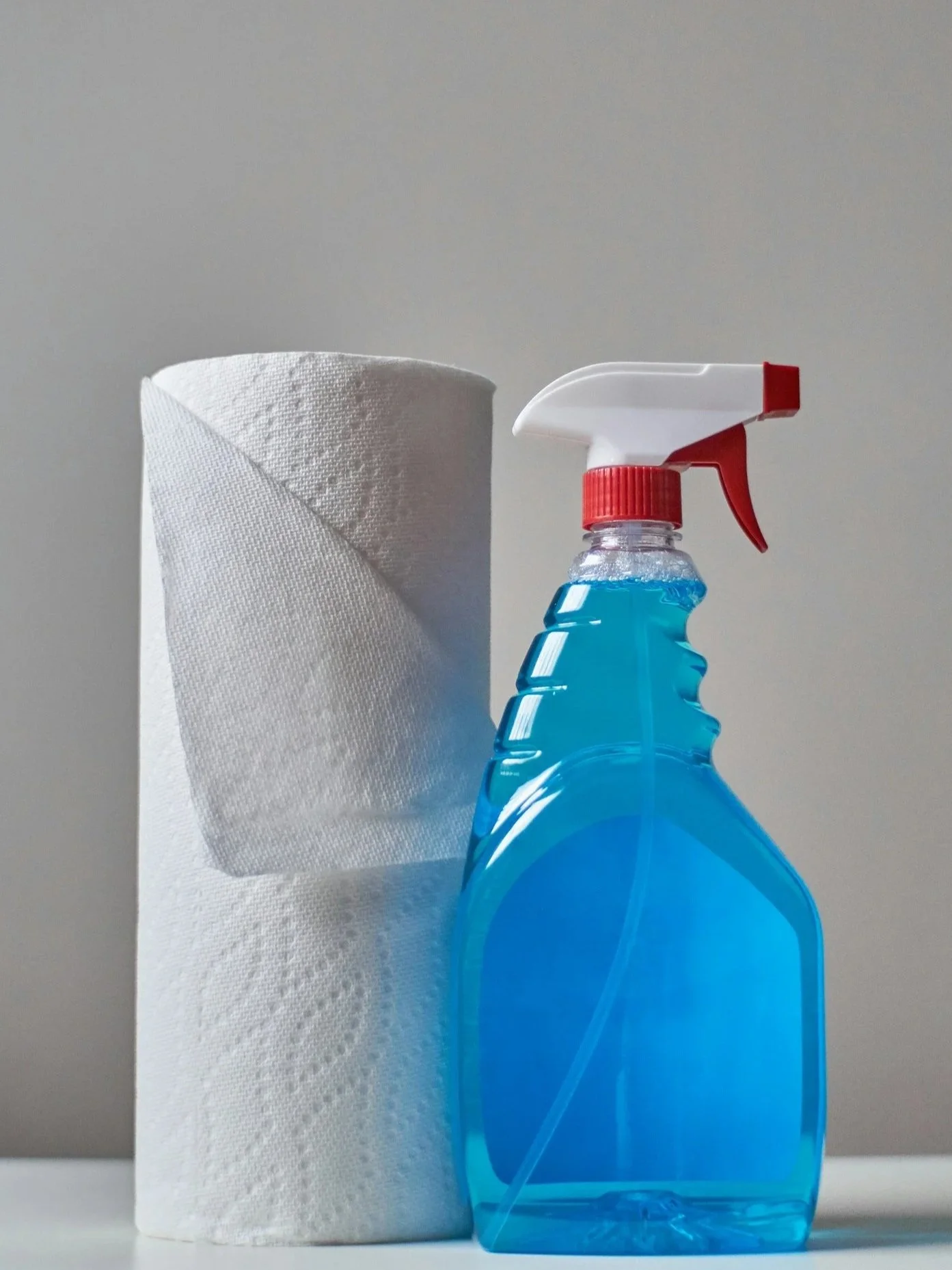 A roll of white paper towels and a spray bottle filled with blue cleaning solution on a white surface against a neutral background.