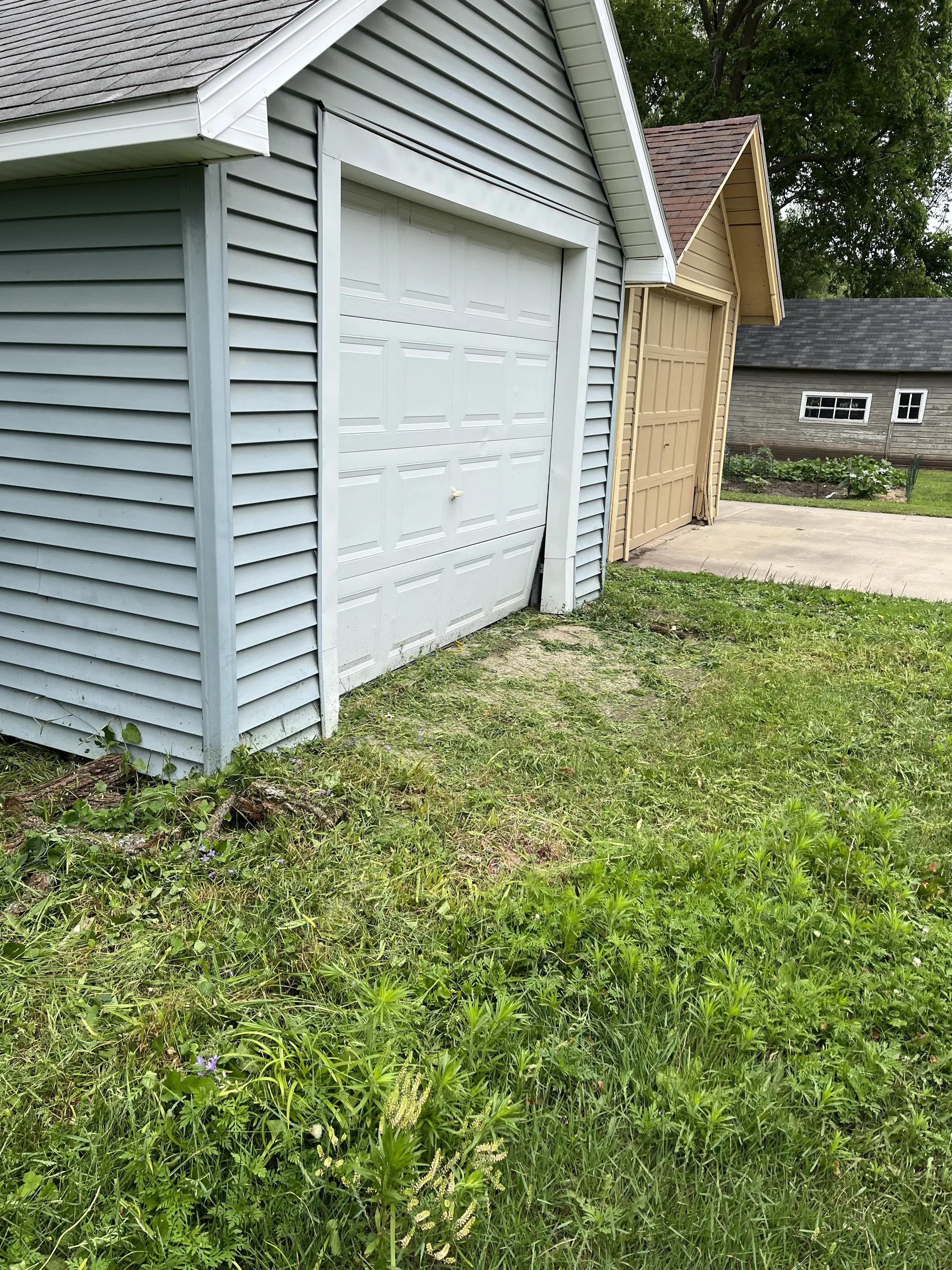 Multiple garages with closed doors in a residential backyard, surrounded by grass, plants, and trees.