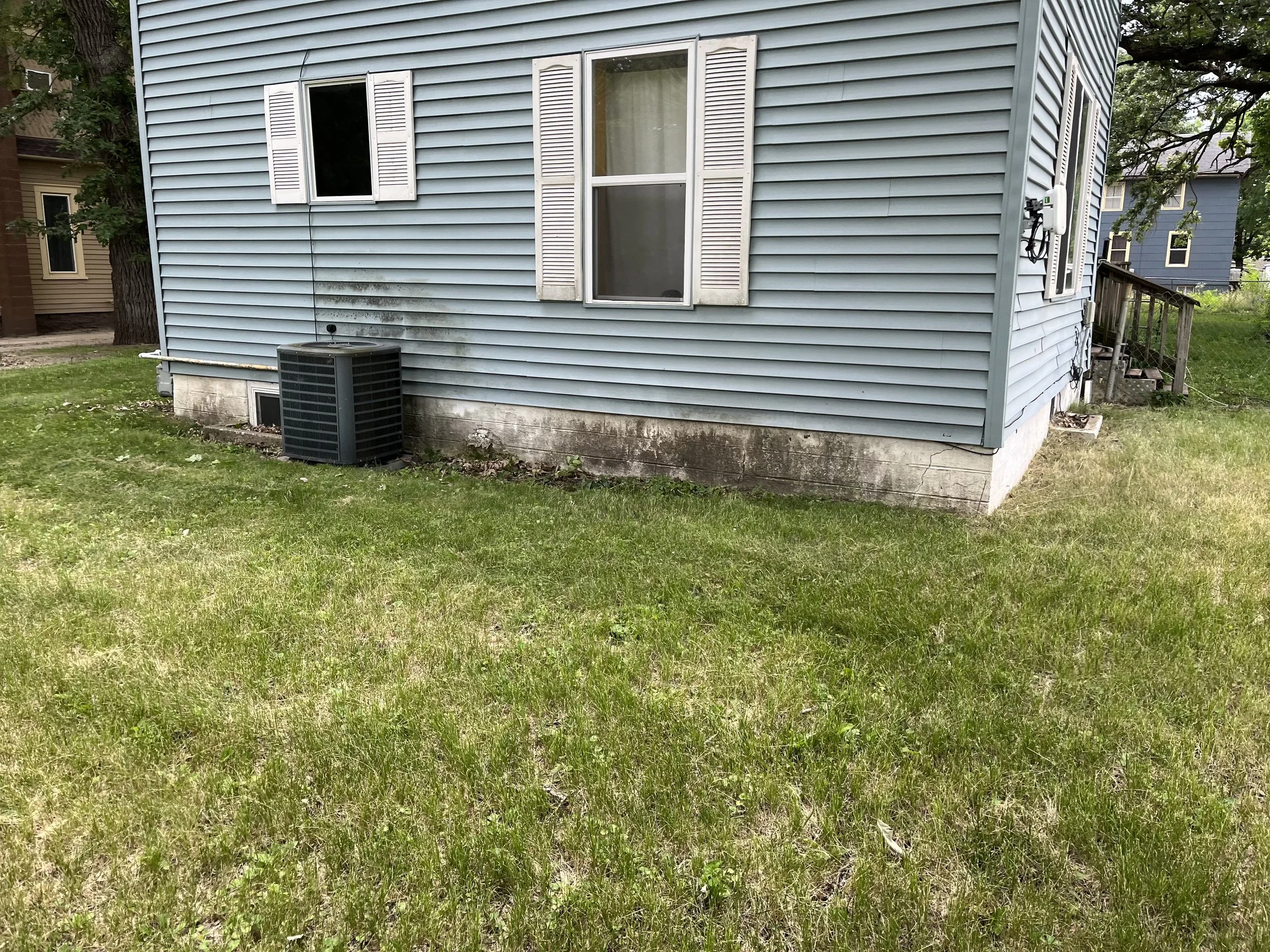 Side view of a light blue house with white shutters, a small basement window, an air conditioning unit, and a grassy yard with some dirt and weeds.