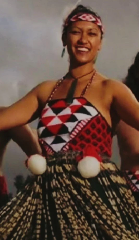 Smiling woman in traditional Māori attire, wearing a patterned dress with red, black, and white geometric designs, a headband, and accessories, outdoors with a cloudy sky in the background.