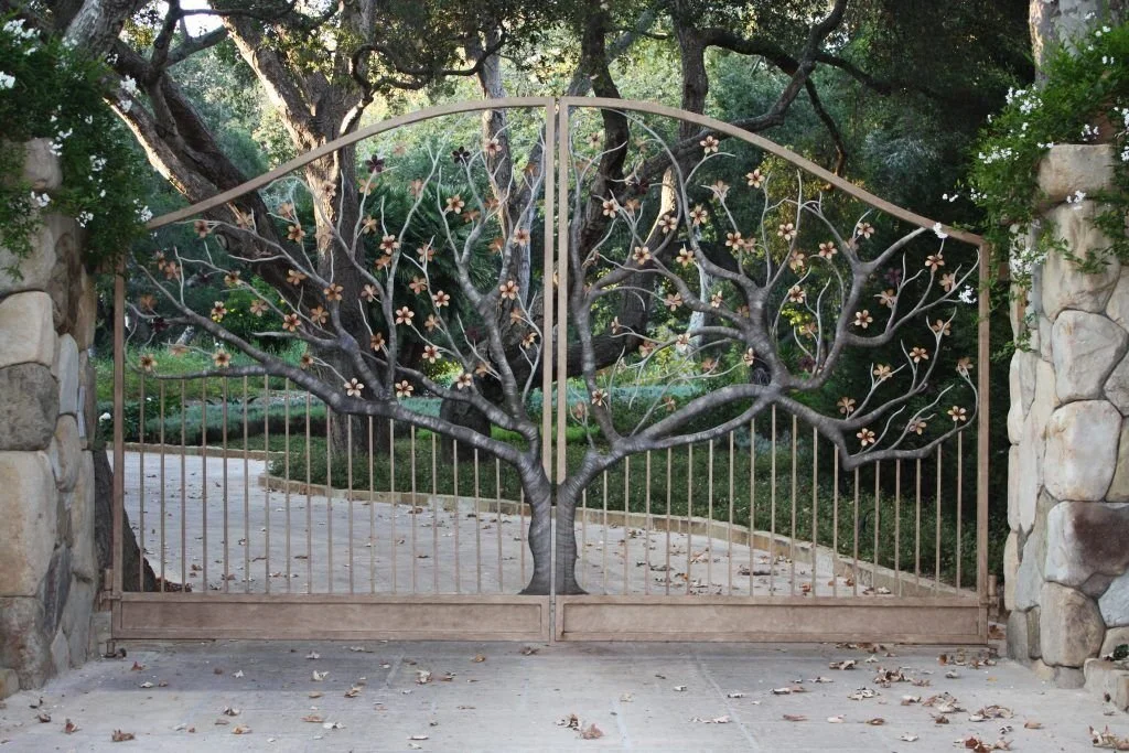 An artistic, handmade wrought iron driveway gate featuring an intricate tree design, complete with a central trunk, sprawling branches, and small metallic flowers, set between two large stone pillars.