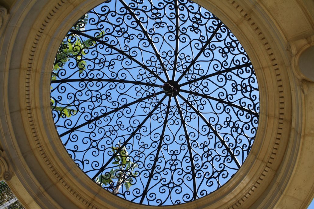 A looking-up view of an ornate black wrought iron dome, featuring intricate radial scrollwork and set within a circular carved stone frame, with a clear blue sky visible through the open metalwork.