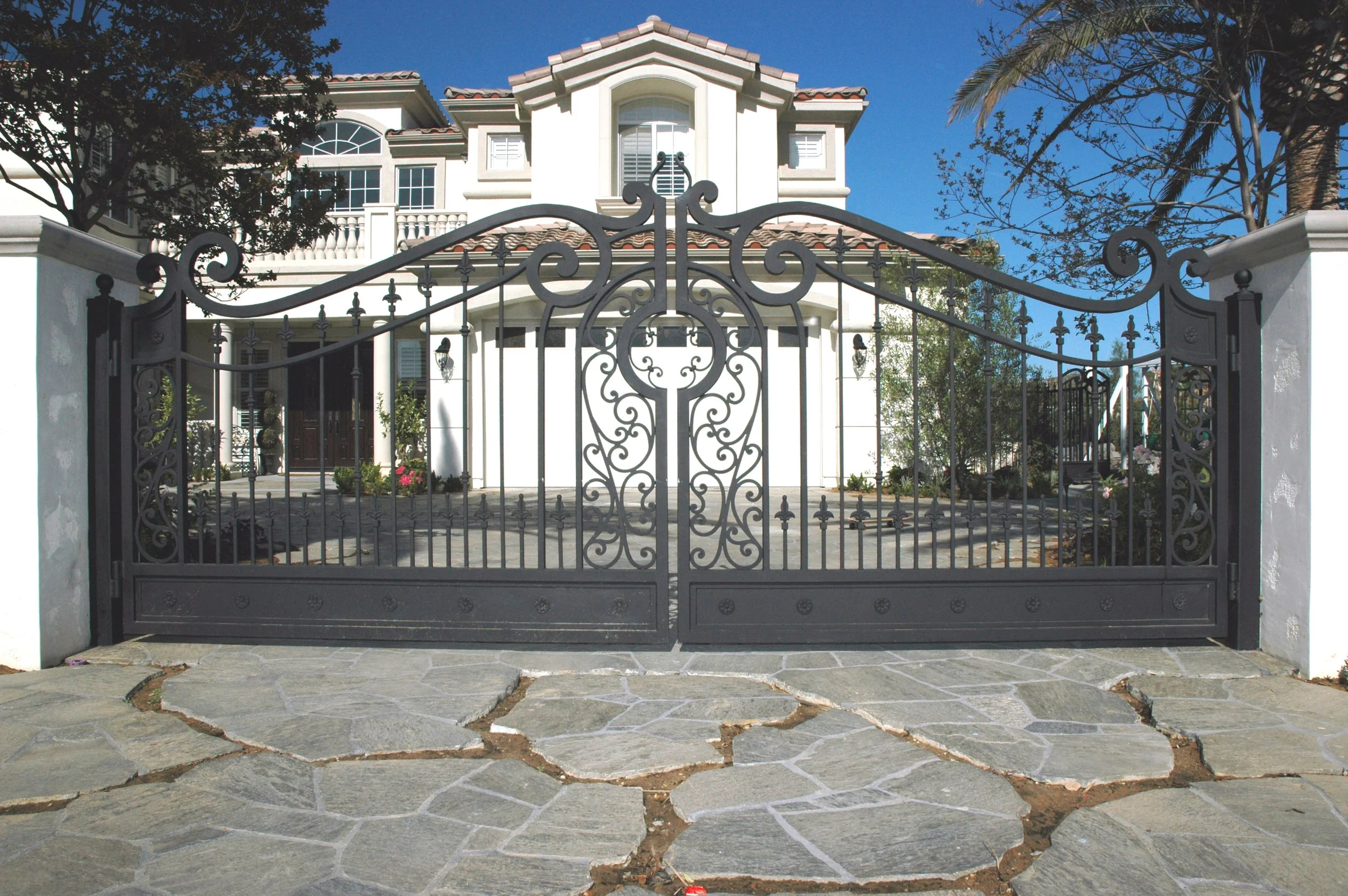 A wrought iron gate in front of a large white house with red tile roof, with trees and a clear blue sky in the background.