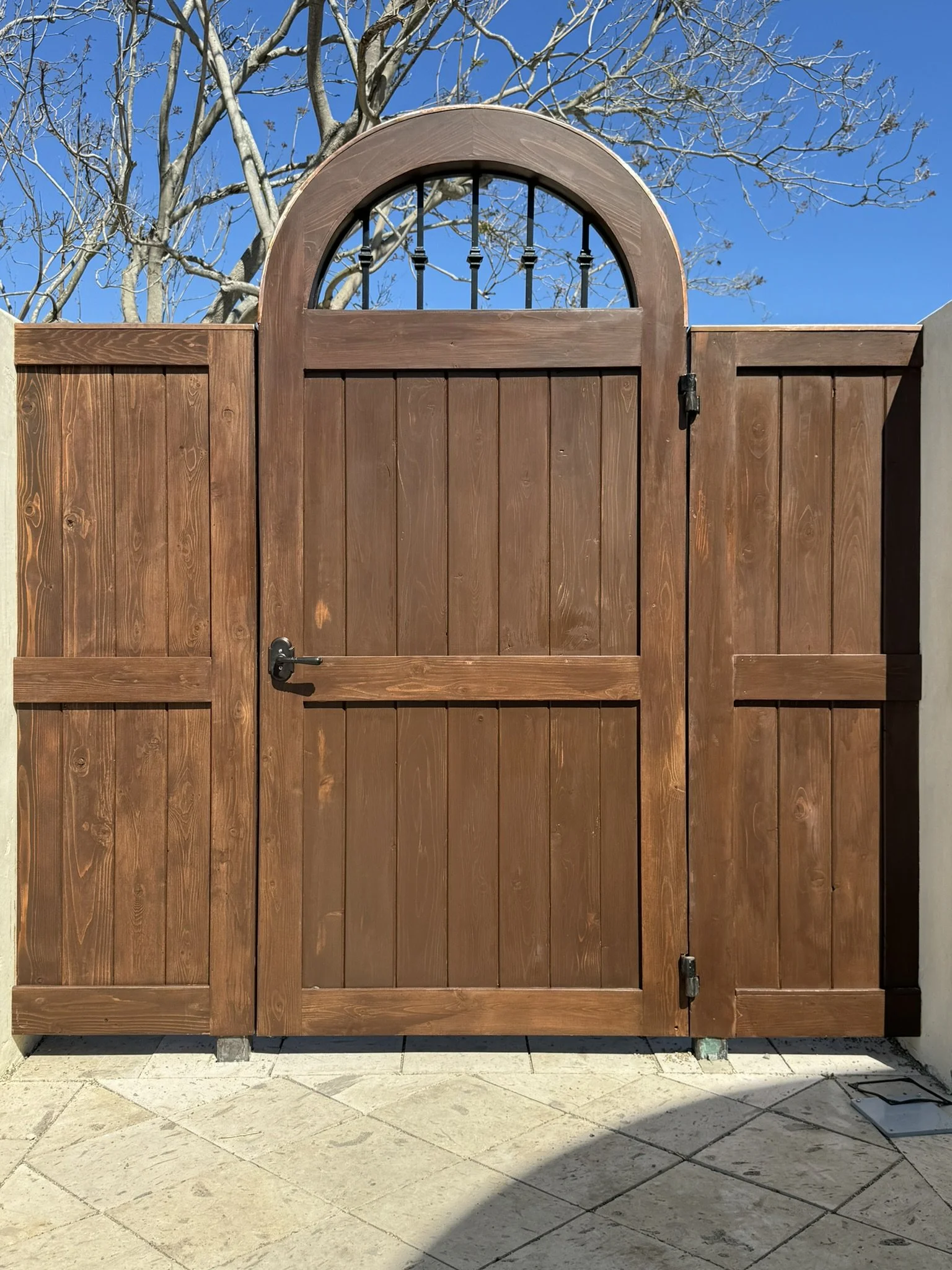 A wooden gate with an arched top and decorative black metal bars, set against a background of bare tree branches and a bright blue sky.
