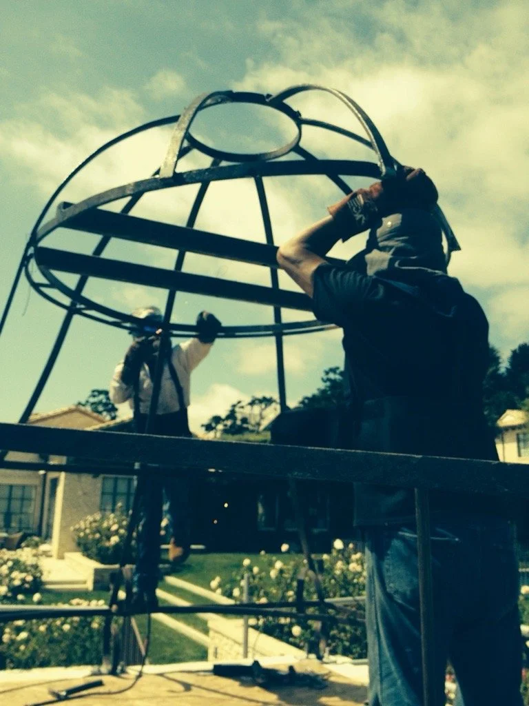 Two workers installing or repairing a large outdoor bell housing on a metal structure in a residential area with houses and flowers.