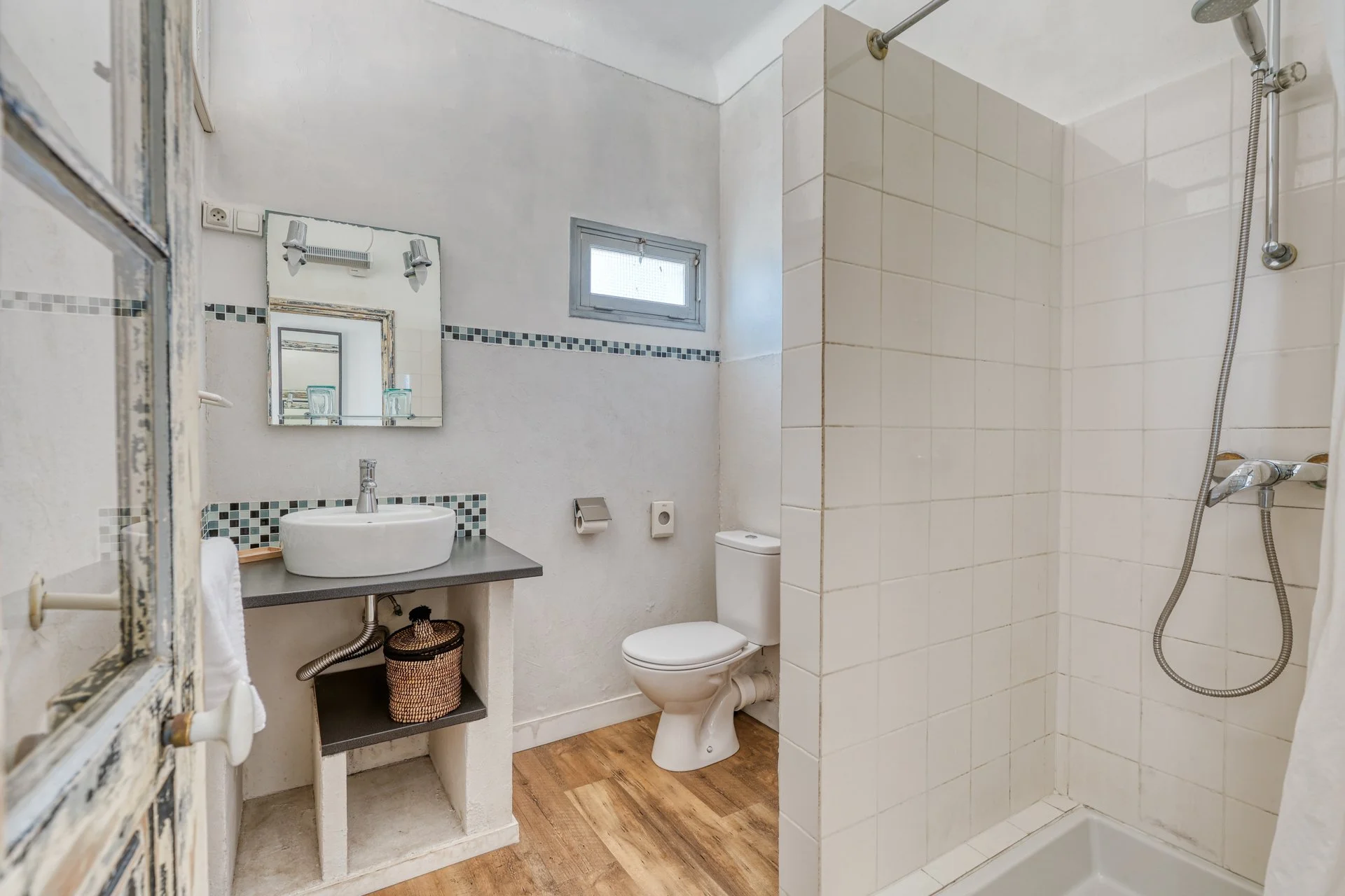 A minimalist bathroom with a small window, a round vessel sink on a black countertop, a mirror, a toilet, and a shower area with white tile walls and a handheld showerhead.