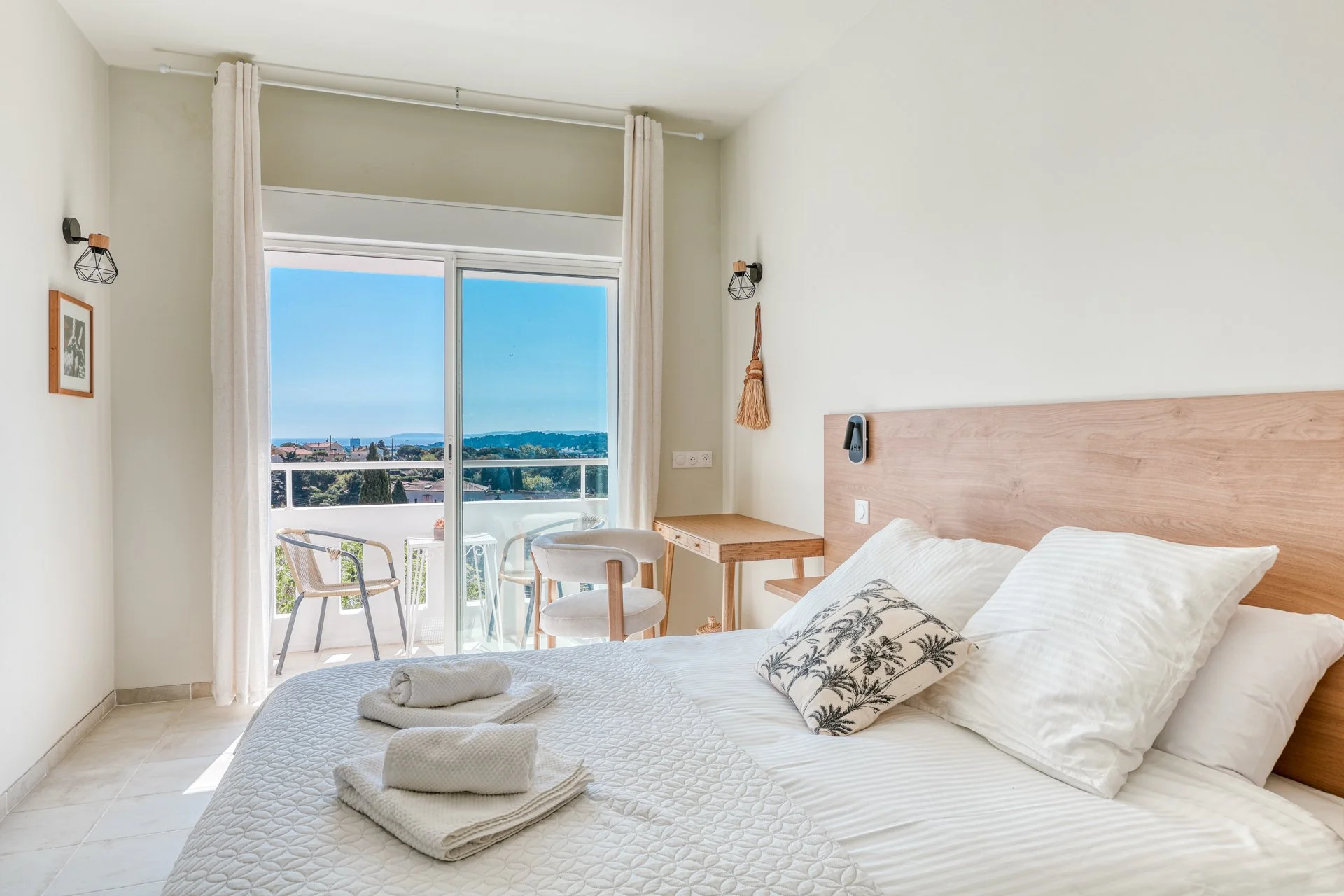 Bedroom with a bed, pillows, and towels, large window with a balcony overlooking a city view, small table and chairs outside, light-colored walls and furniture, and natural sunlight.