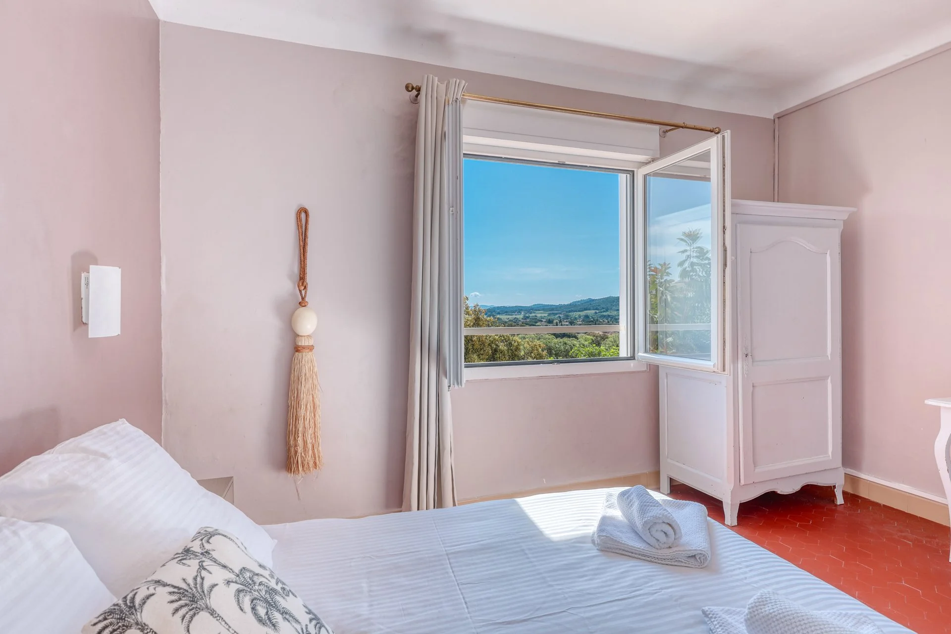 A bedroom with a large open window showing a hilly area with houses and trees, a palm tree, and a partly cloudy blue sky. The room has a bed with white bedding, a wooden headboard, a small shelf, and a modern black wall-mounted device. There's a white towel on the bed.