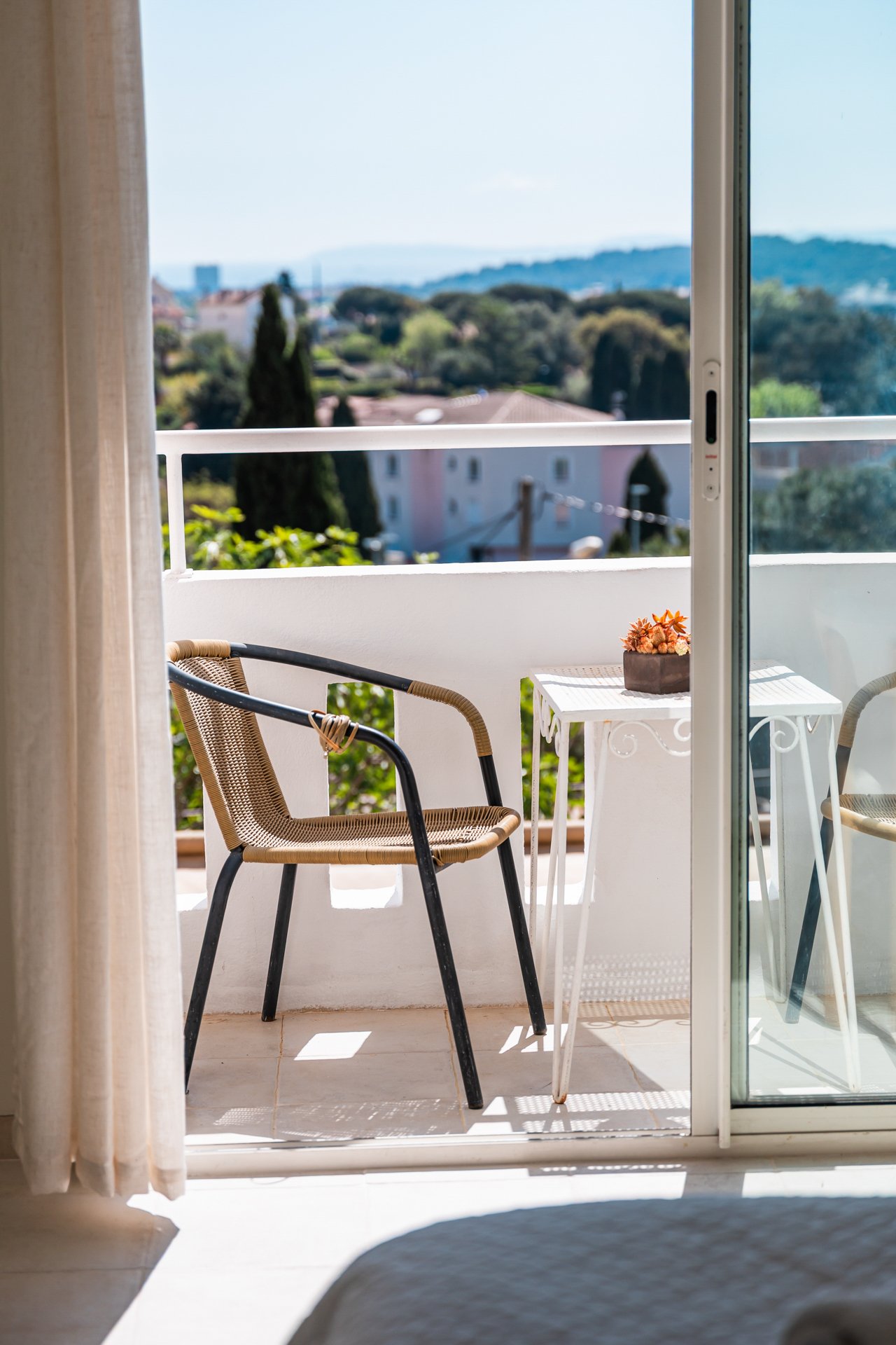 Balcony with two chairs, a small white table with a plant on it, overlooking a residential neighborhood with trees and hills in the background.