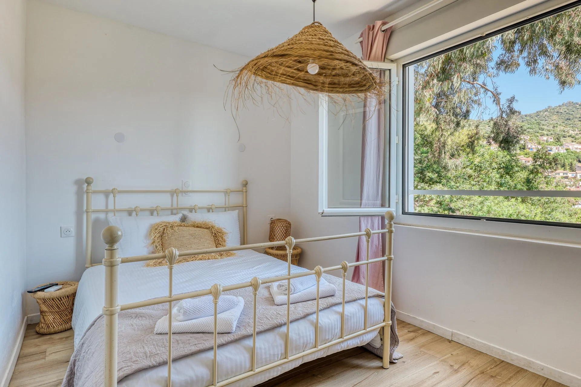 A cozy bedroom with a cream-colored metal bed frame, white bedding, and a textured pillow. Two wicker baskets sit beside the bed, with one holding a remote. A large window with pink curtains is open, showing a view of green trees and distant hillside houses. A rustic woven pendant light hangs from the ceiling.