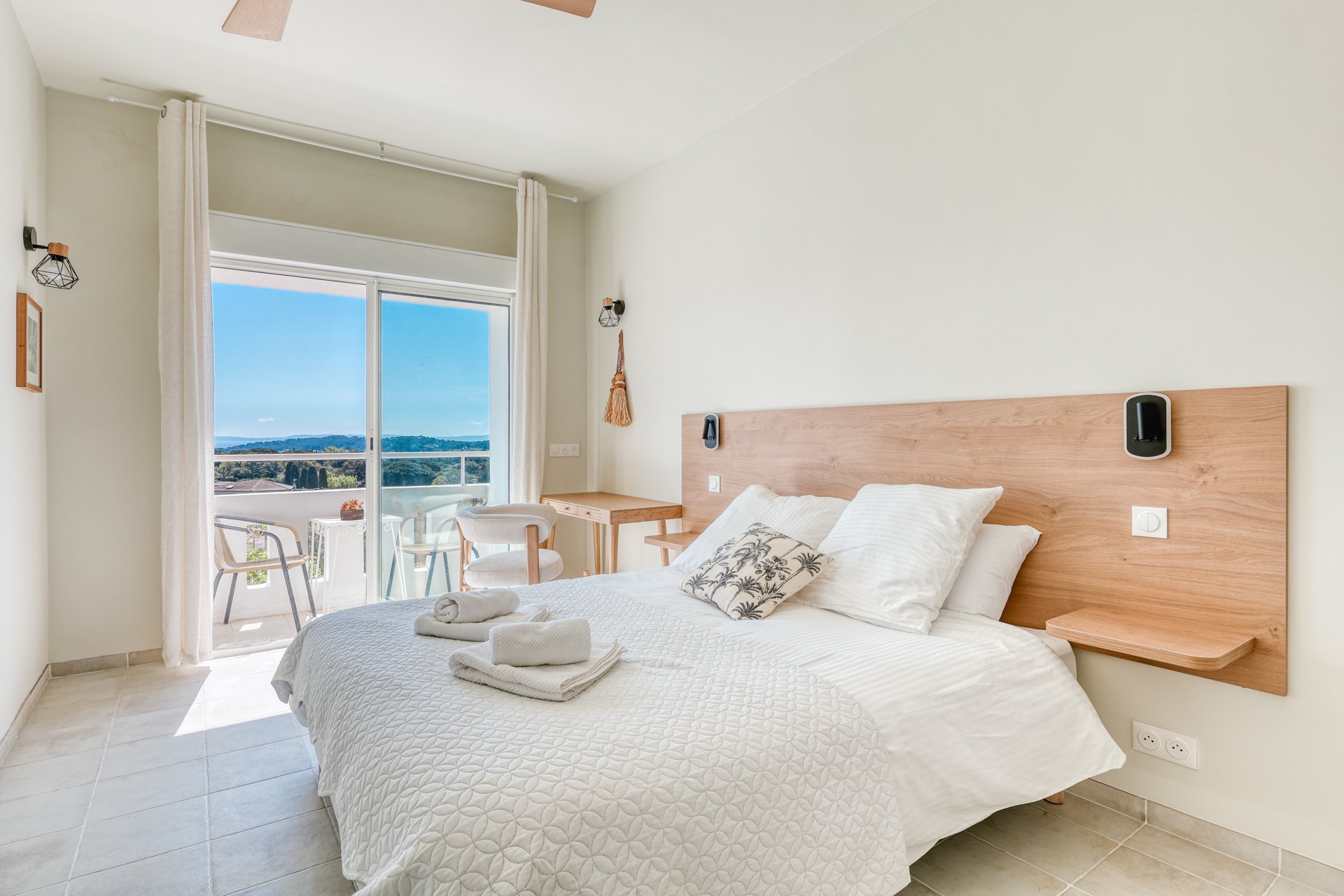 Bright hotel room with big bed, wooden headboard, white bedding, and towels. Balcony with outdoor chairs and scenic view of trees and hills.