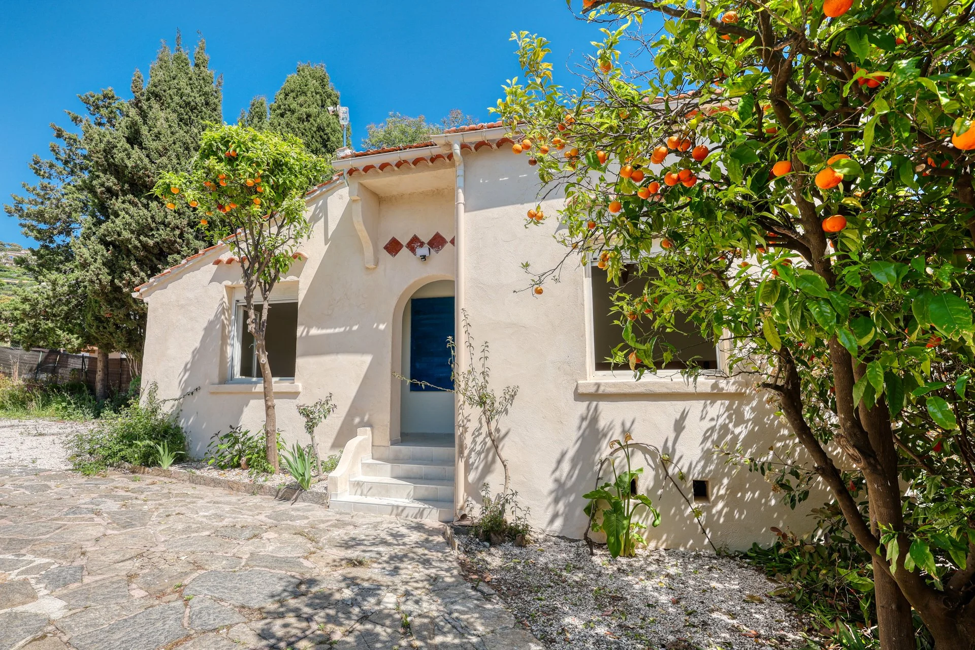 White house with an arched entrance and steps, surrounded by orange trees with ripe oranges, on a sunny day with a clear blue sky.