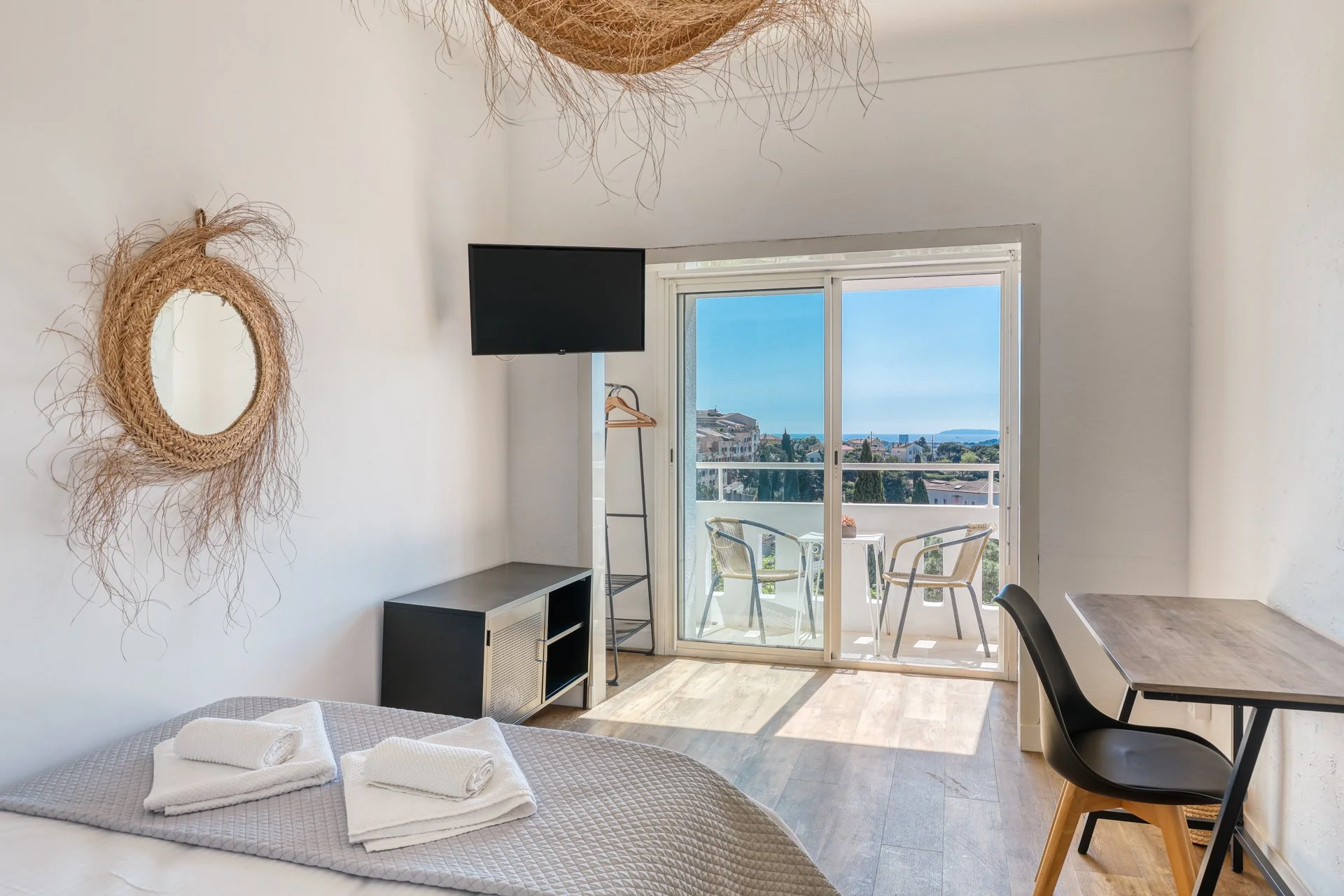 Interior of a bright apartment with a bed, towels, a small TV, a sliding glass door leading to a balcony with outdoor chairs, a view of rooftops and the sea, and decorative wall mirrors.