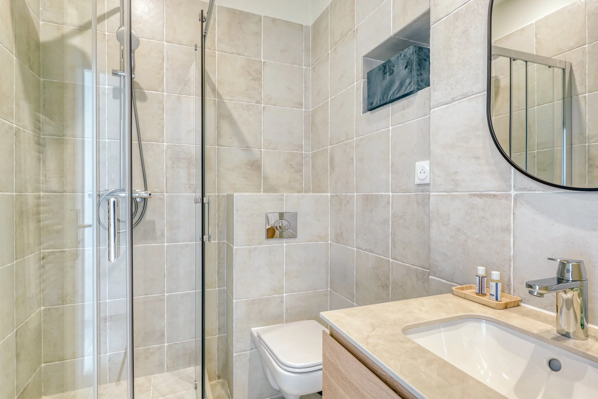 Modern bathroom with beige tiled walls, glass shower enclosure, round mirror, bathroom sink, beige countertop, and some toiletry bottles on a wooden tray.