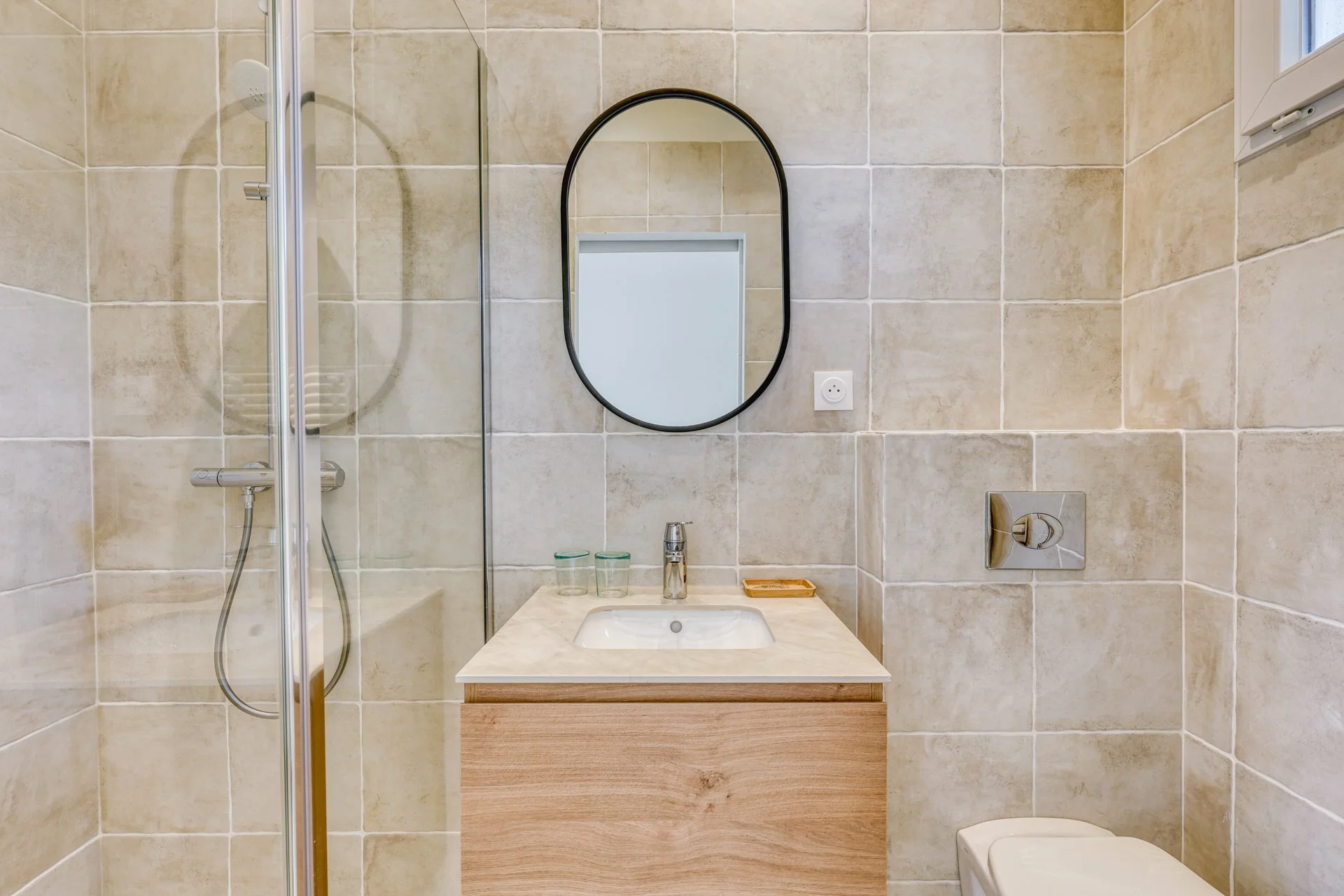 A modern bathroom with beige tiled walls, a shower with a glass door, a small sink with a wooden cabinet, a round mirror with a black frame, and a toilet. Two glasses and a soap tray are on the sink.