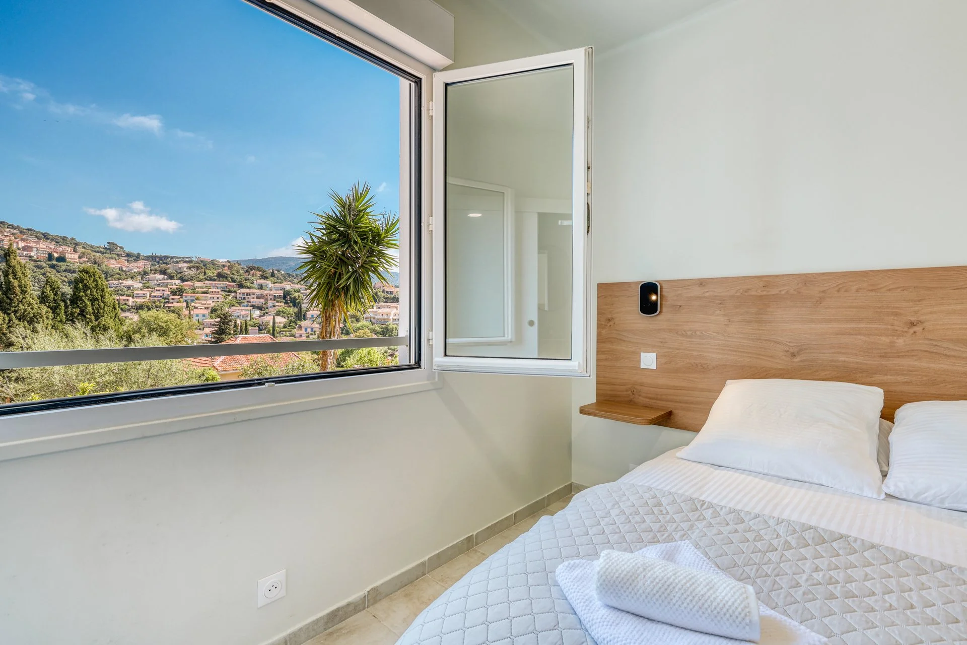 A bedroom with a large open window showing a hilly area with houses and trees, a palm tree, and a partly cloudy blue sky. The room has a bed with white bedding, a wooden headboard, a small shelf, and a modern black wall-mounted device. There's a white towel on the bed.