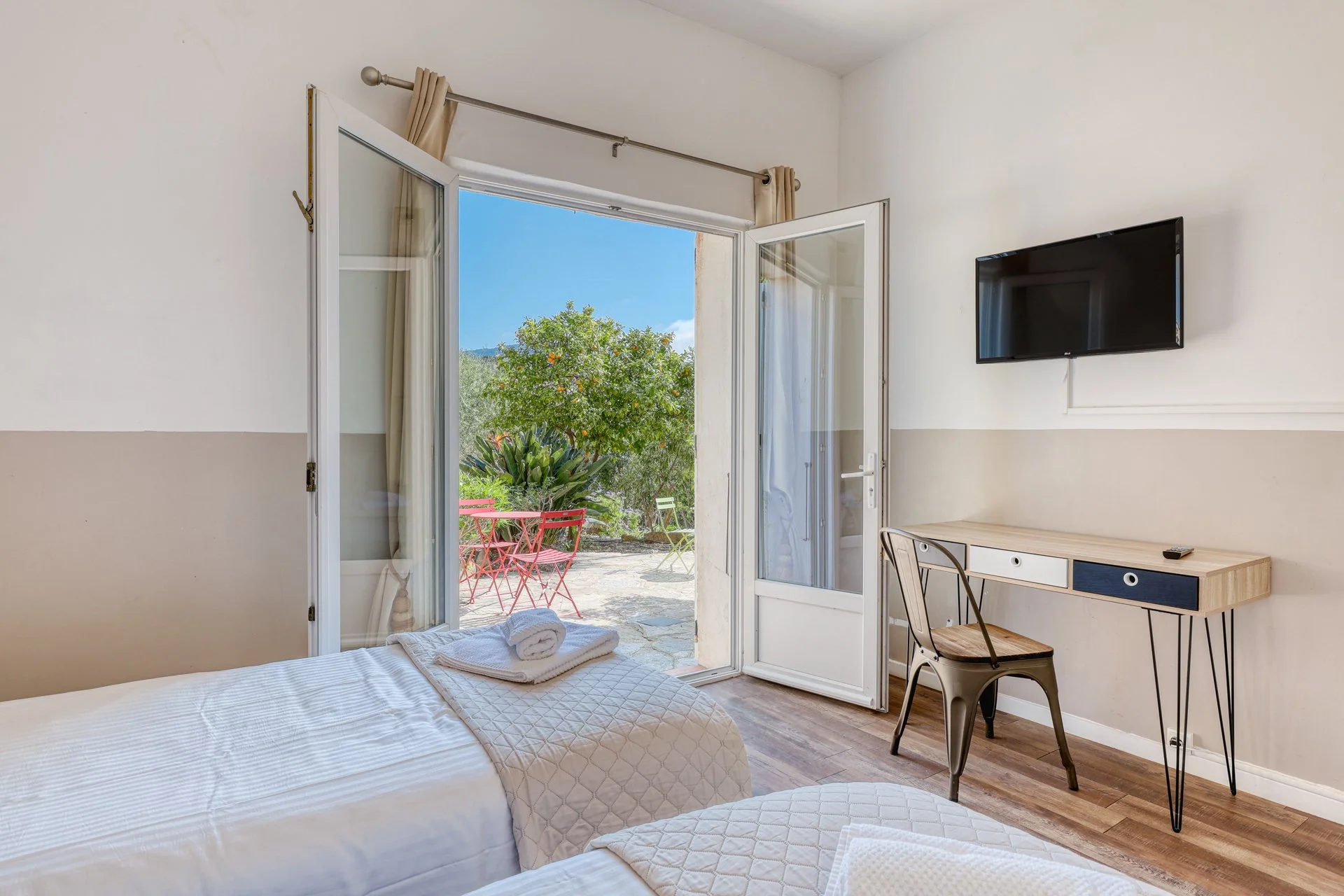 A bright hotel room with an open door leading to a garden patio with red and green outdoor furniture and lush greenery.