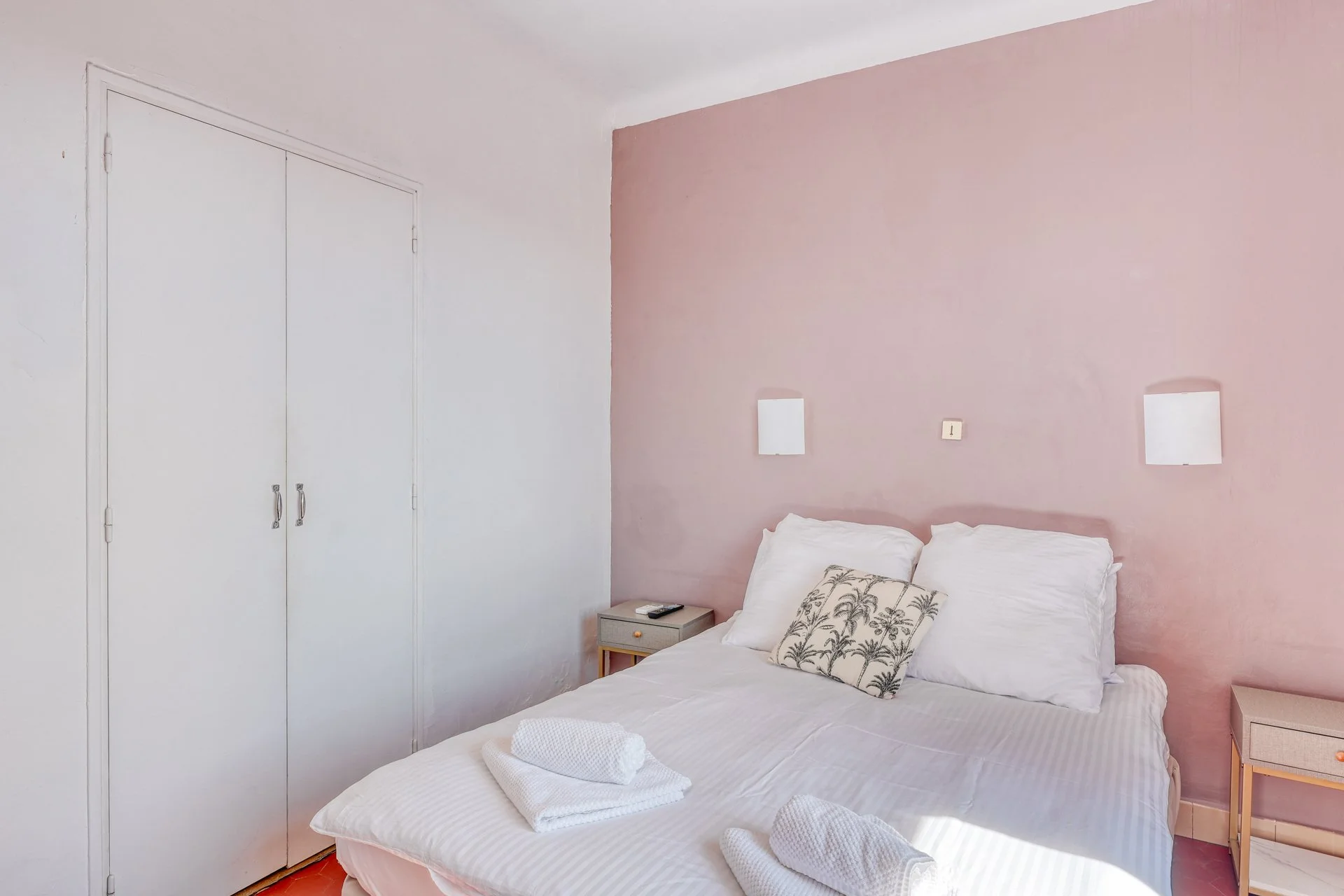 Bedroom with a white bed, pink accent wall, two white wall-mounted lamps, a small gray nightstand with a phone, towel, and pillow with palm tree print.