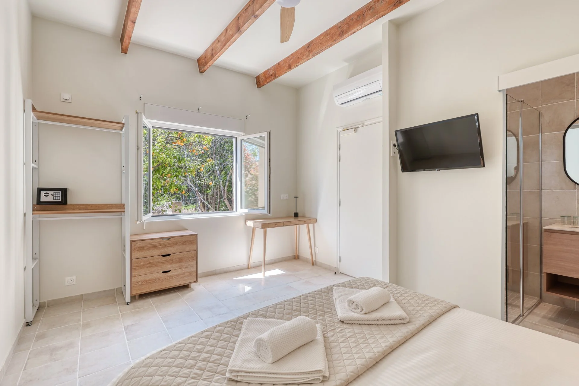 A bright, minimalist bedroom with a bed, folded towels, a small wooden dresser, a window with green trees outside, a wall-mounted flat-screen TV, and wooden ceiling beams.