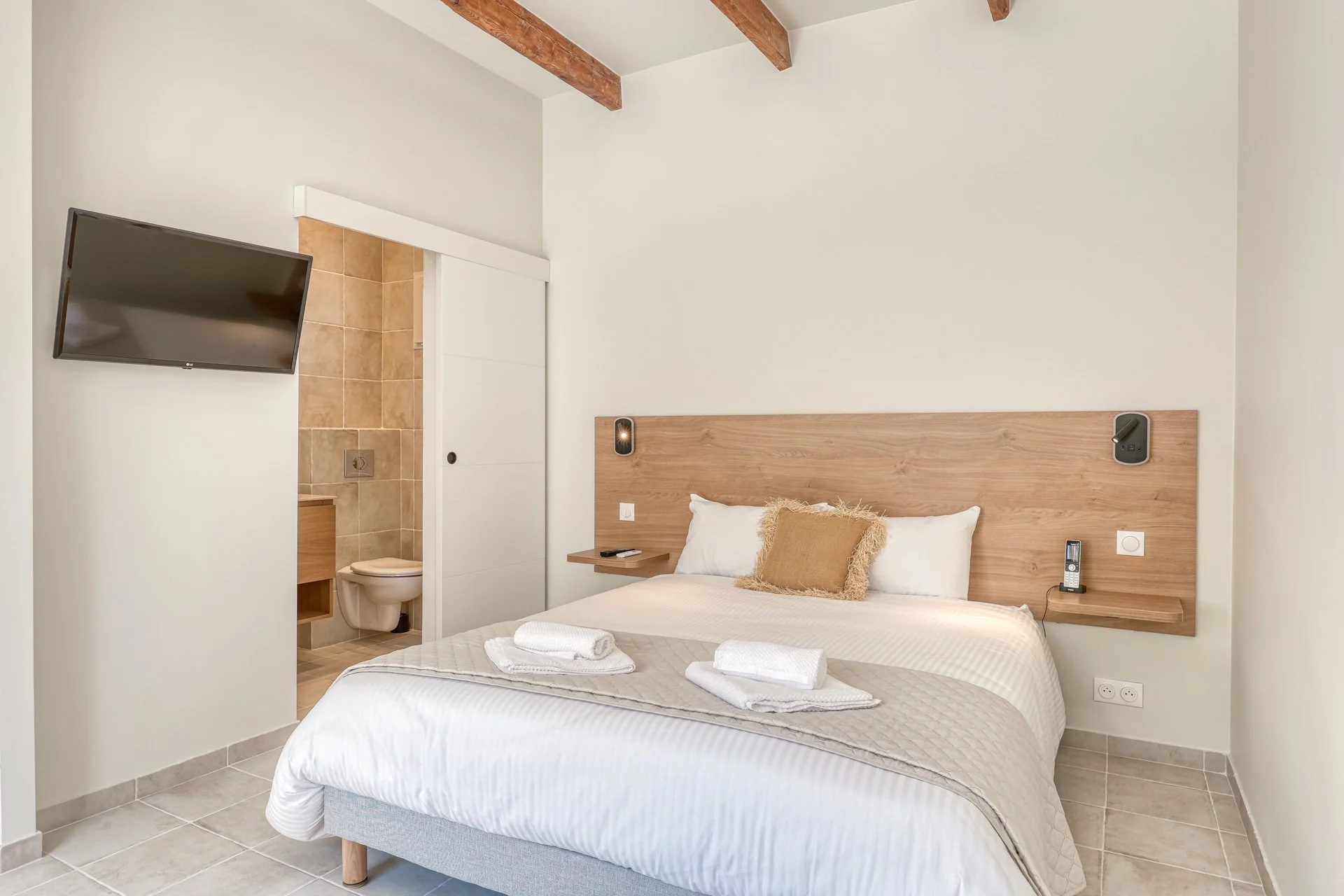 Bedroom with a bed, pillows, and towels, large window with a balcony overlooking a city view, small table and chairs outside, light-colored walls and furniture, and natural sunlight.