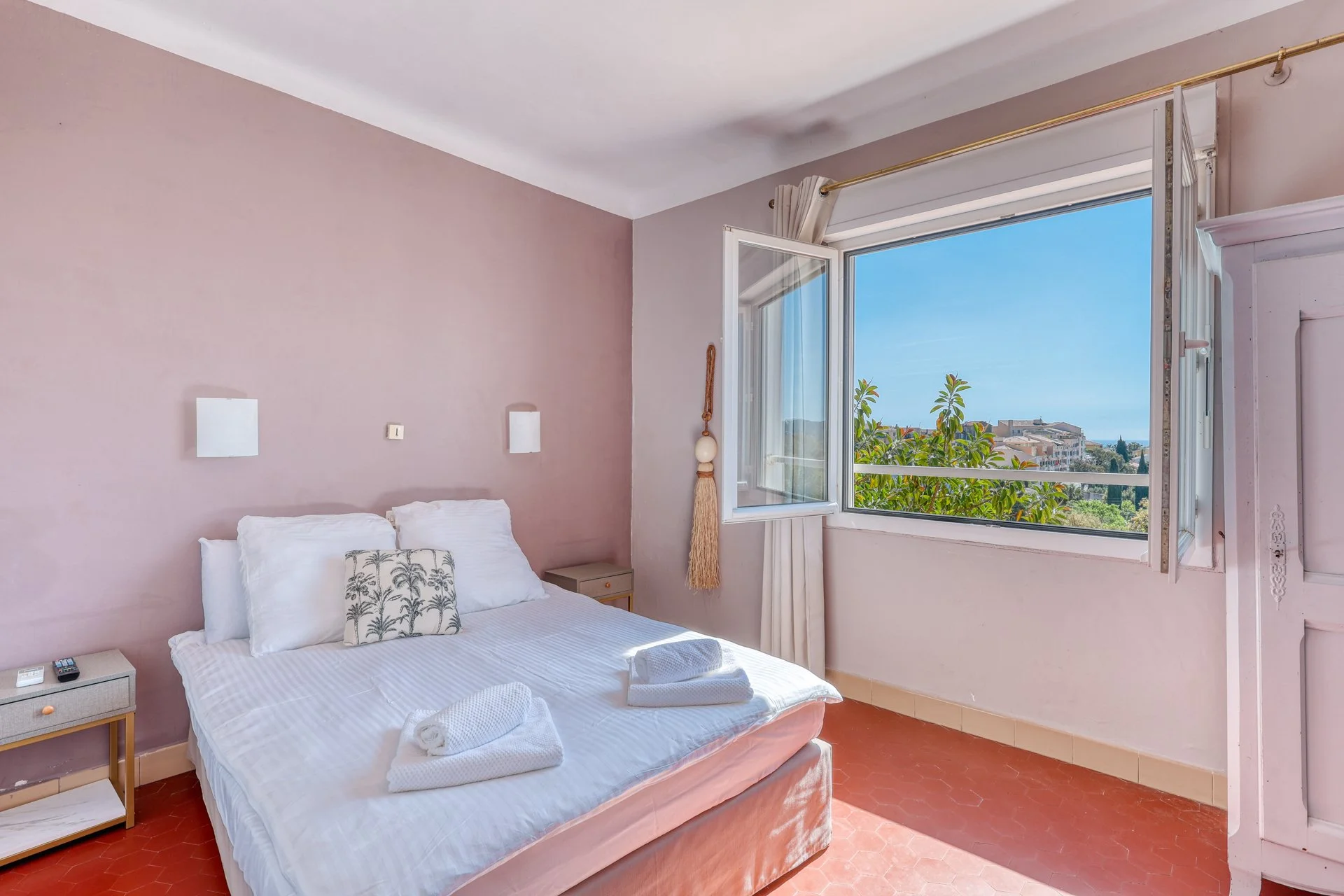 A bedroom with a bed, bedside table, and decorative pillows, open window showing a view of greenery and buildings under a blue sky.