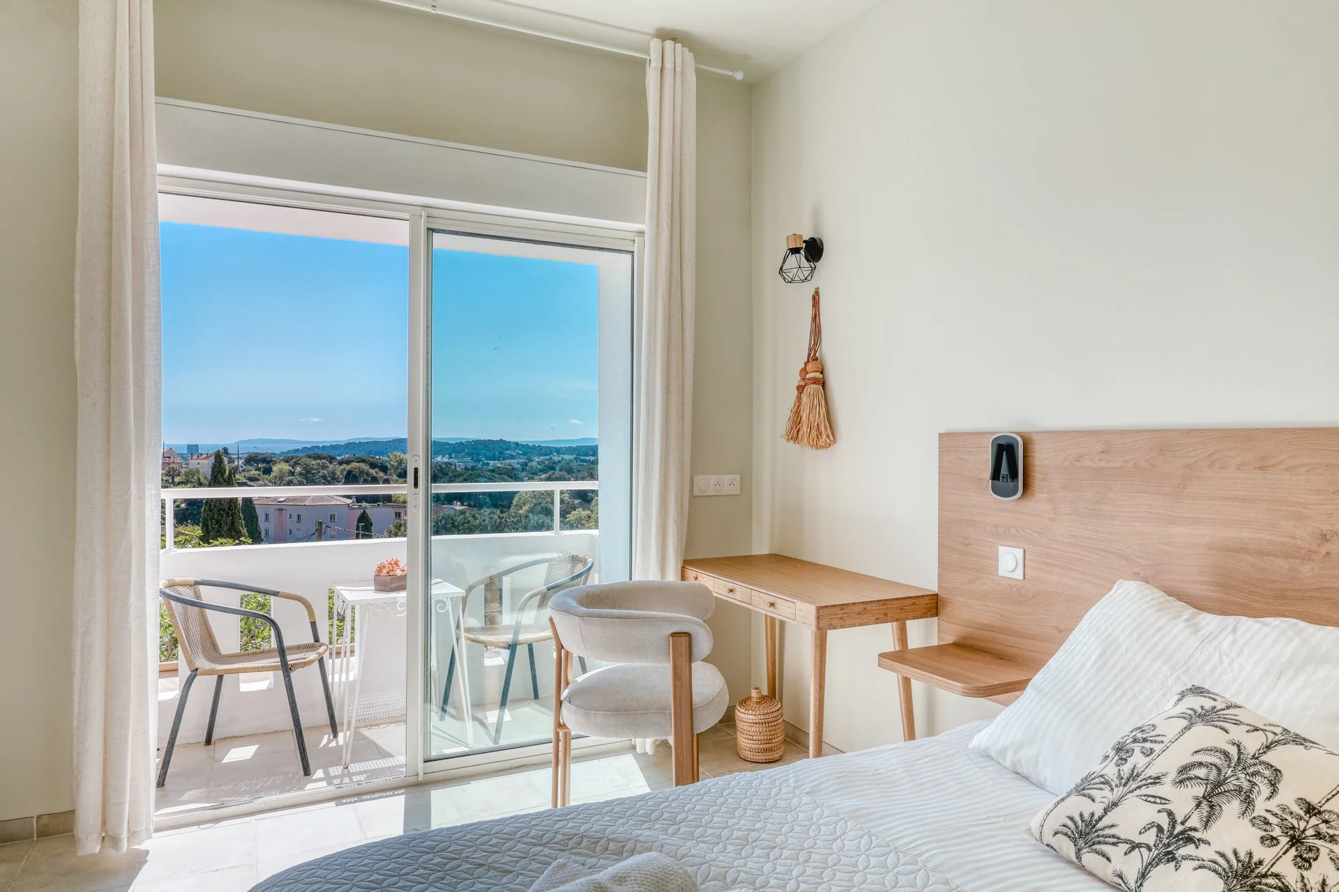 Hotel room with bed, wooden headboard, small desk, beige chair, balcony with outdoor seating, view of trees and distant hills, light-colored walls, window with curtains.