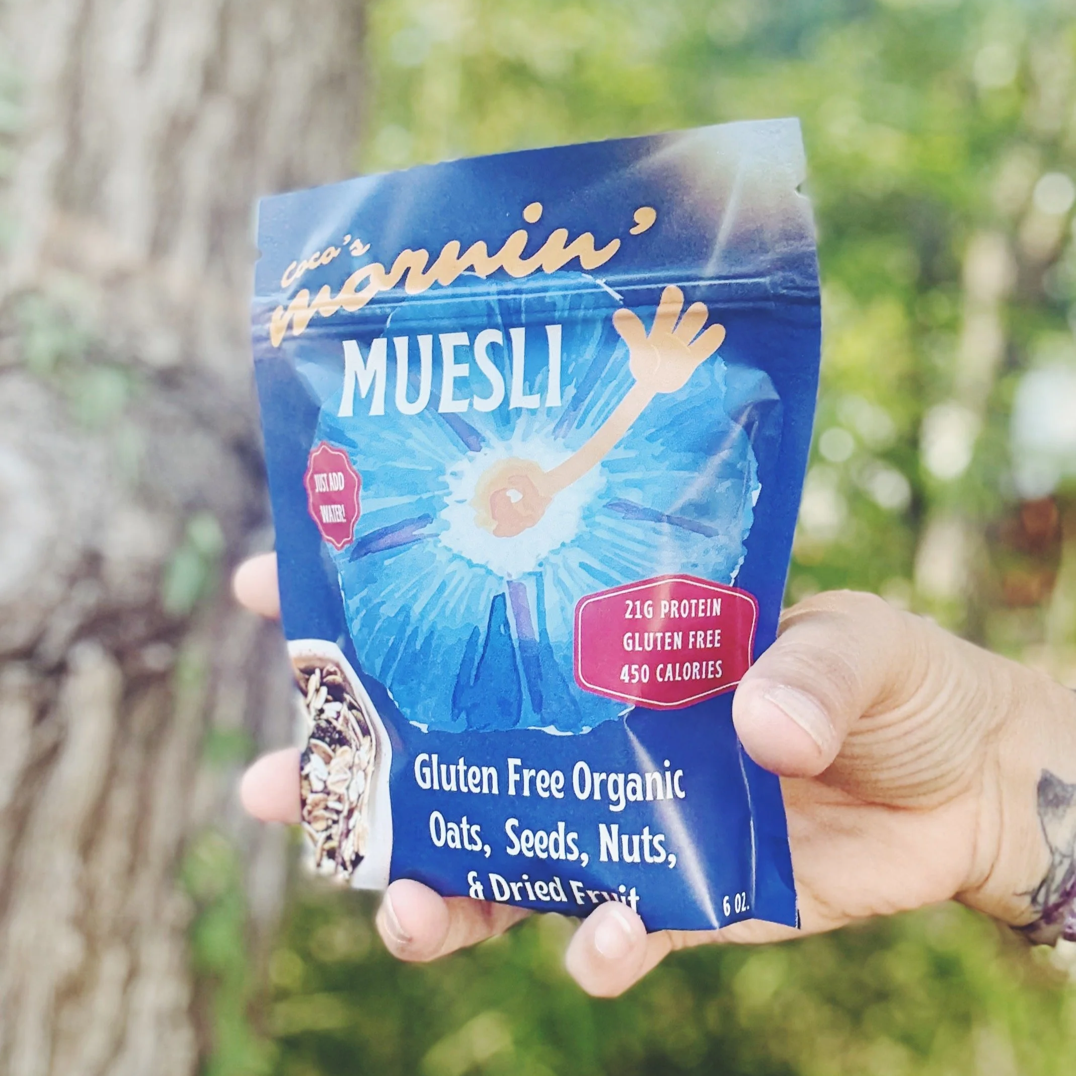 Hand holding a blue bag of organic gluten-free oatmeal muesli outdoors with trees in the background.