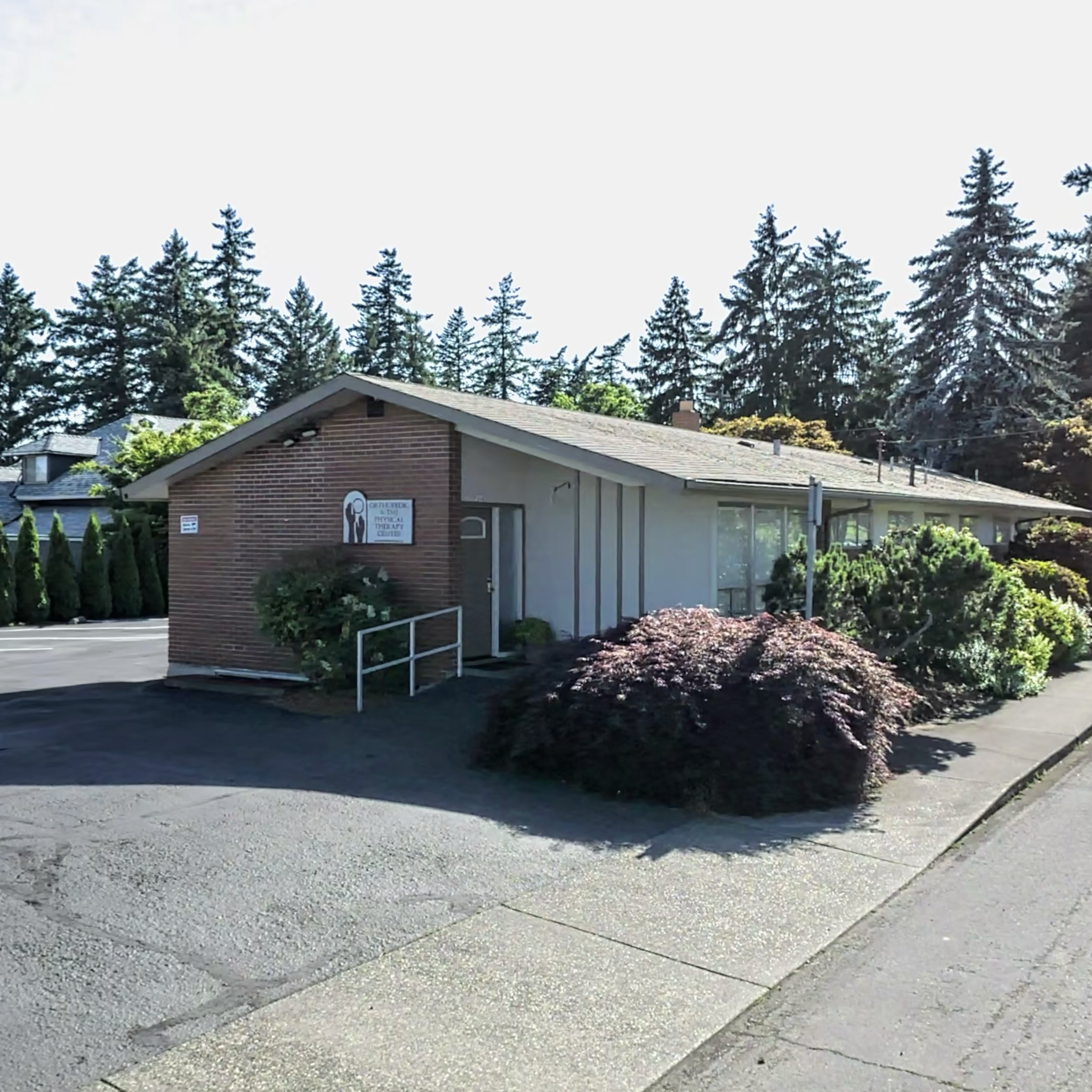 Exterior view of a small medical or therapy clinic building with a brick facade on one side and light gray siding on the other, surrounded by greenery and trees, with a parking lot and sidewalk in front.
