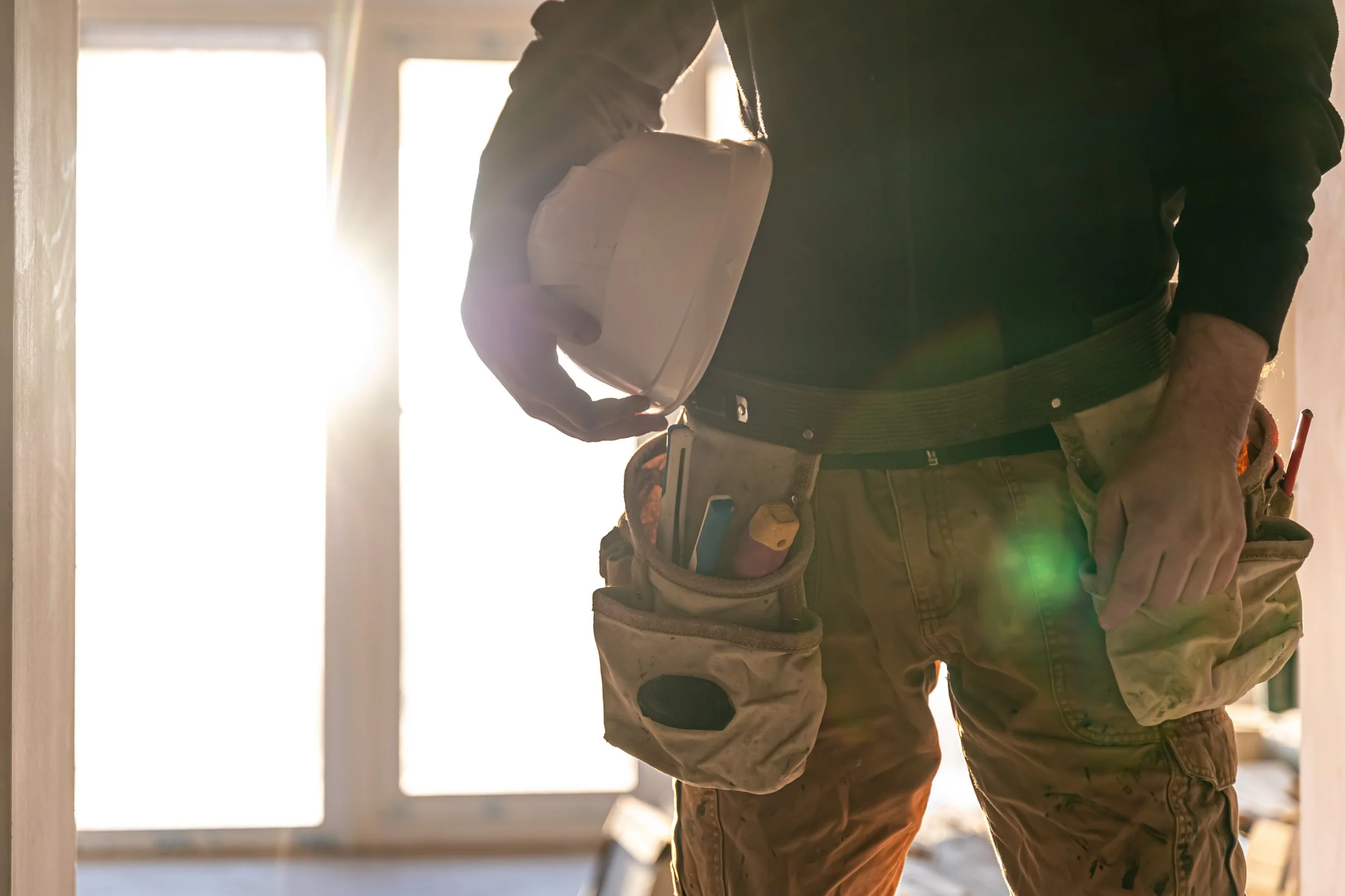 A construction worker wearing a tool belt and holding a white hard hat at a building site with sunlight coming through windows.