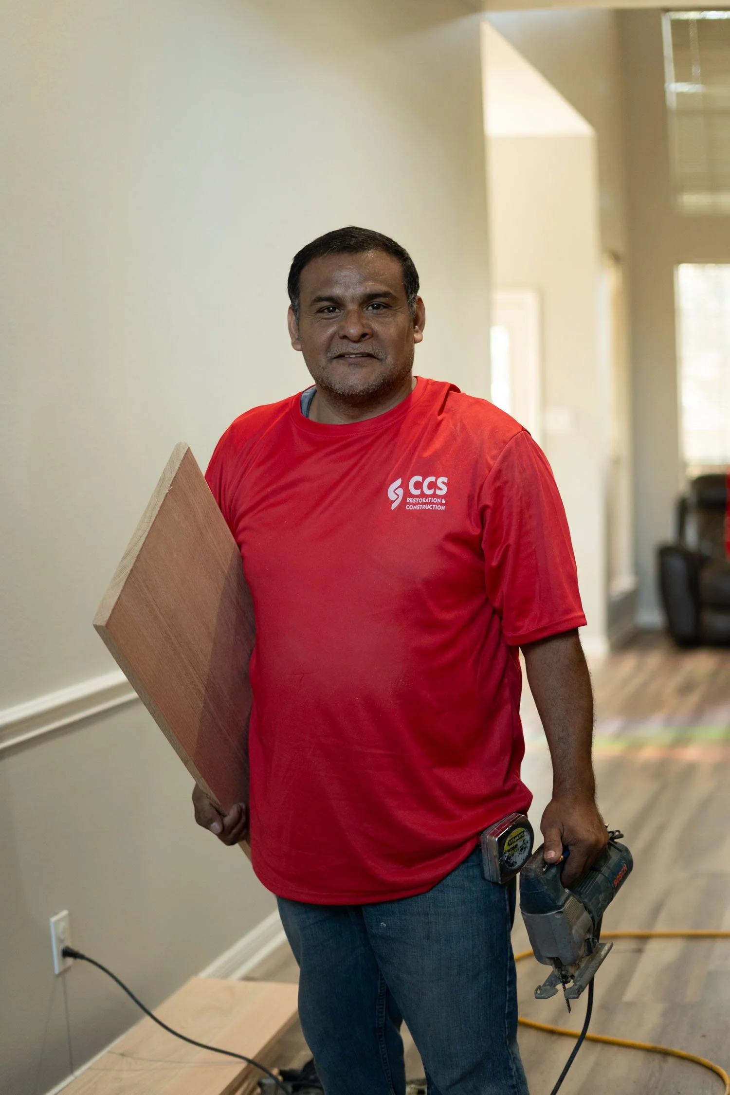 A man in a red CCS Restoration and Construction T-shirt holding a wooden board and a power tool, standing in a room with hardwood floor and light-colored walls.